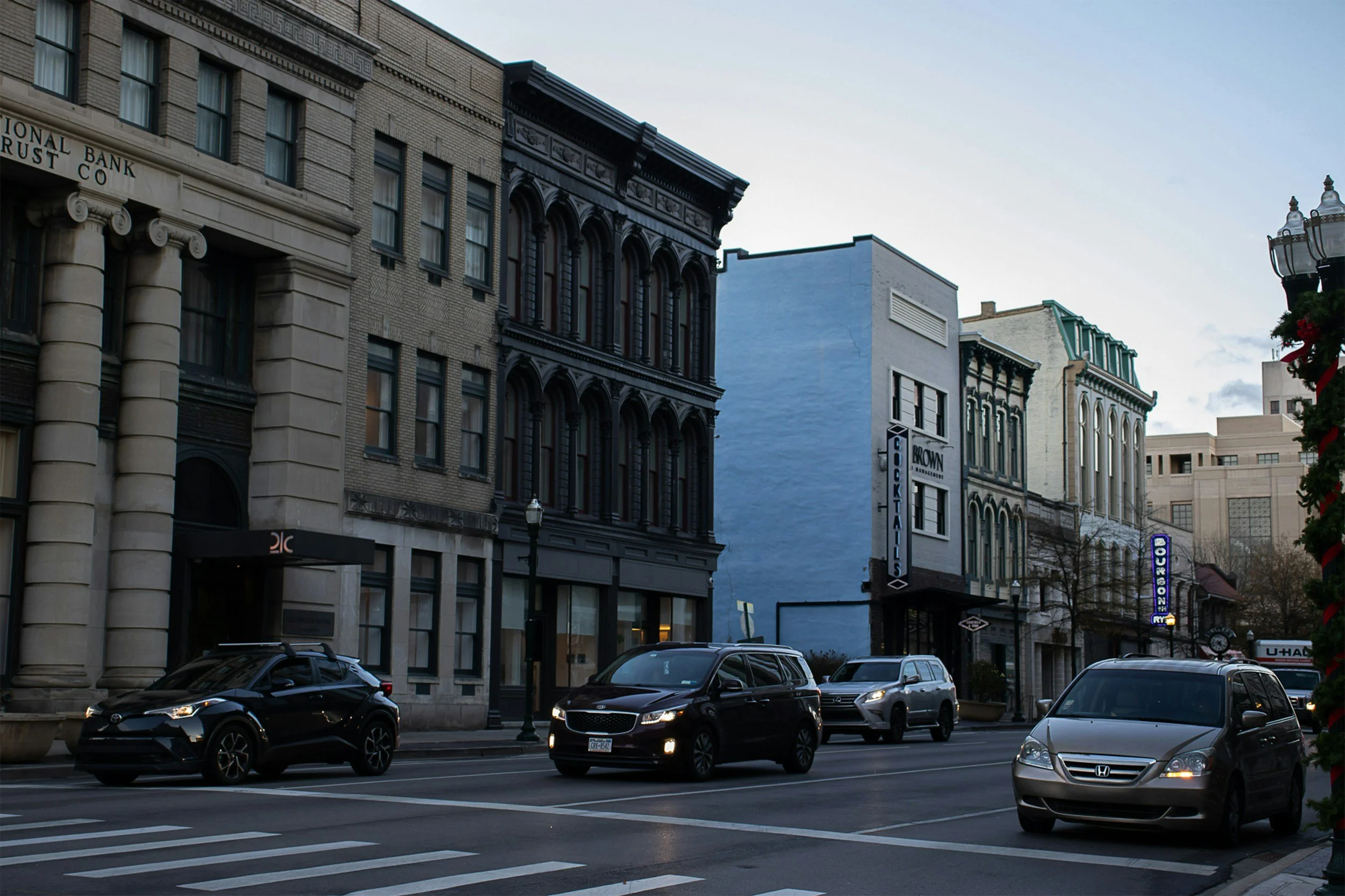 City street with parked cars, historic buildings, and a street lamp against a cloudy sky.