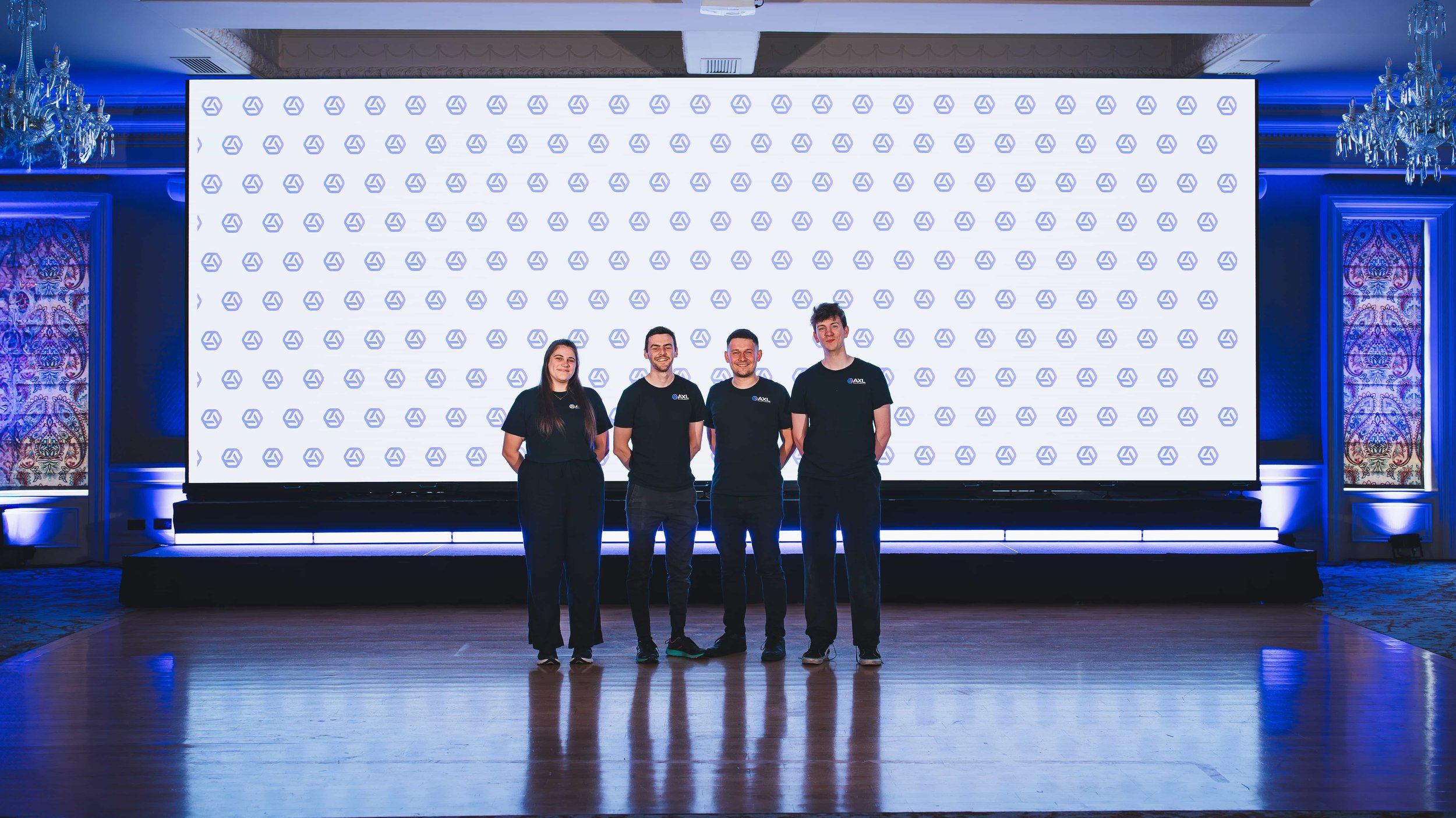 Four people standing in front of a large screen with a logo pattern at an indoor event.