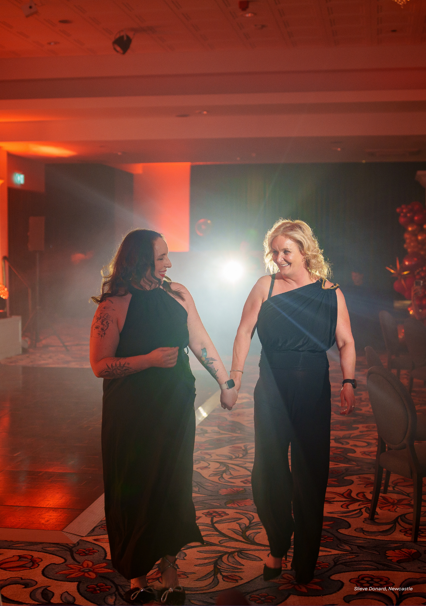 Two women wearing black dresses holding hands and smiling at each other on a dance floor with decorative carpet, dim lighting, and balloons in the background.