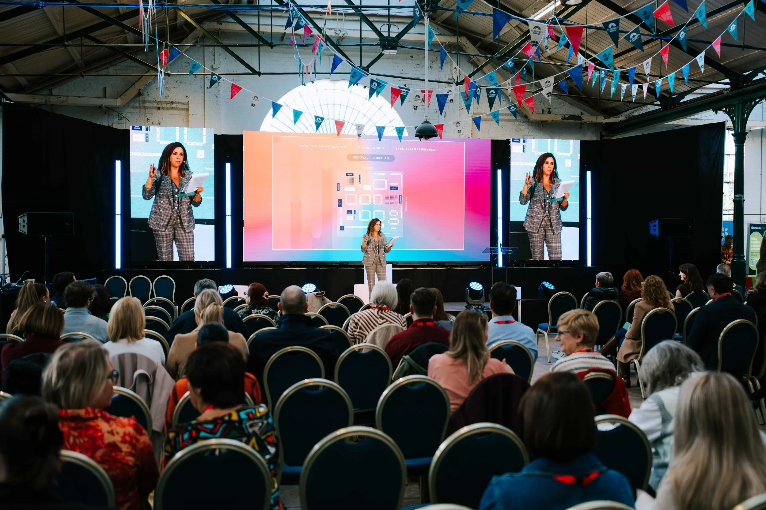 A woman in a gray plaid suit speaking on stage at a conference with a large audience. The stage has two large screens displaying her image, and colorful triangular flags decorate the ceiling. The audience is seated facing the stage in an industrial-style venue with high ceilings and large windows.