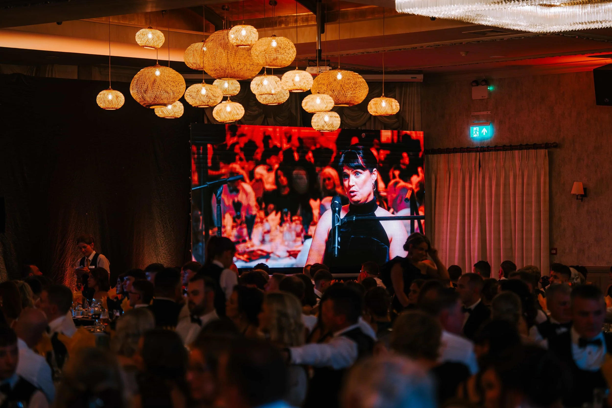 A large crowd attending a formal event, with many people dressed in tuxedos and evening gowns, seated at tables. There is a big screen displaying a woman speaking into a microphone, and hanging decorative lights above.