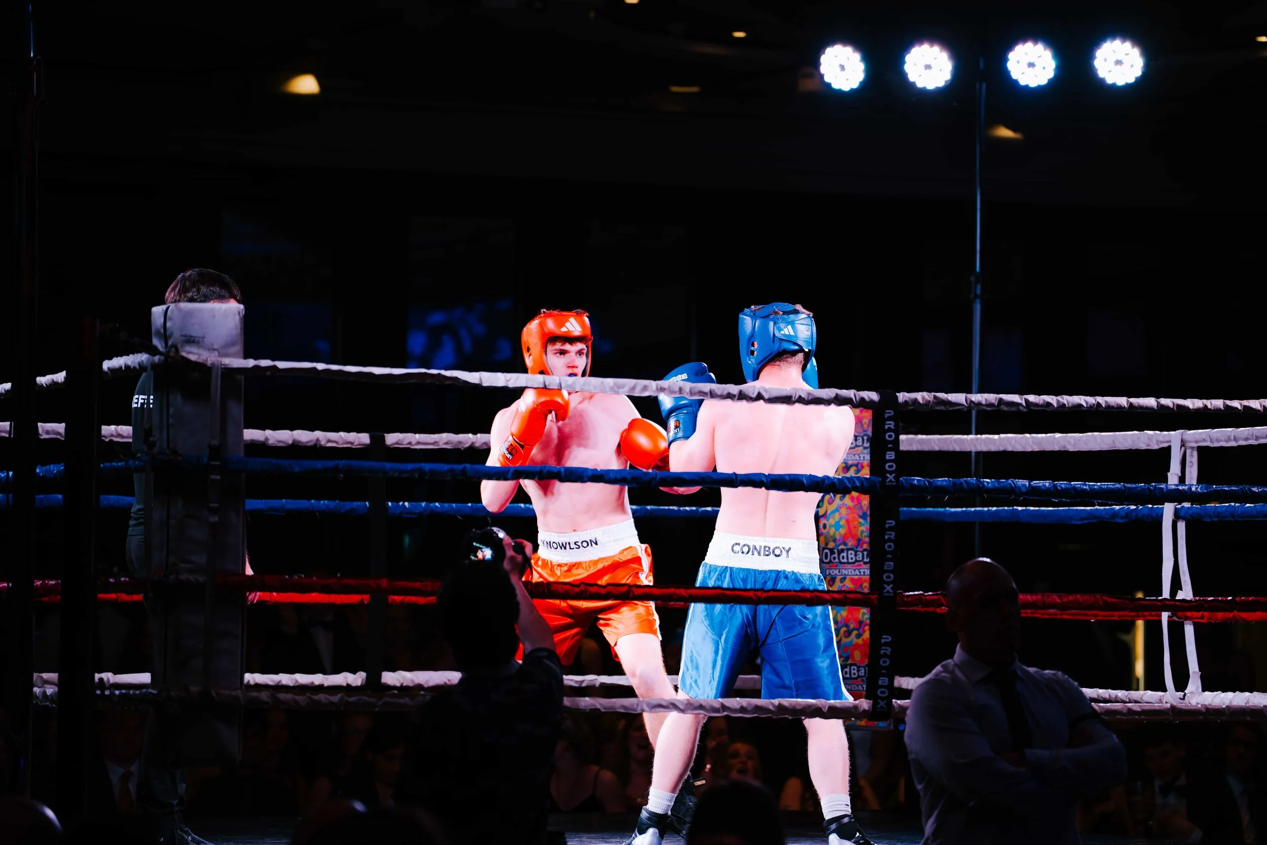 Two young male boxers wearing headgear and gloves sparring in a boxing ring, with a referee observing.