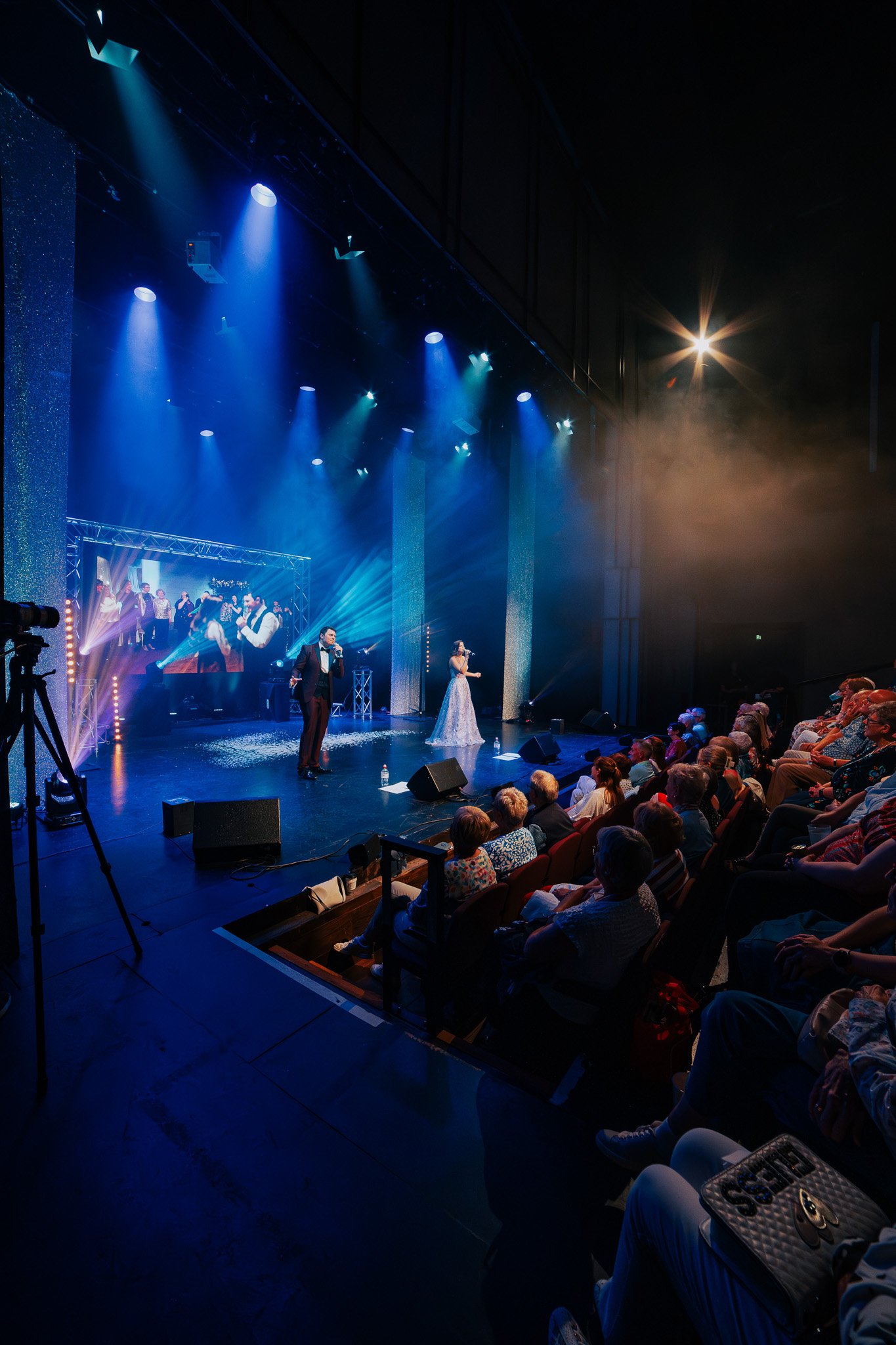 A stage with performers and an audience watching a concert or show, with colorful stage lighting and a large screen displaying the performance in the background.