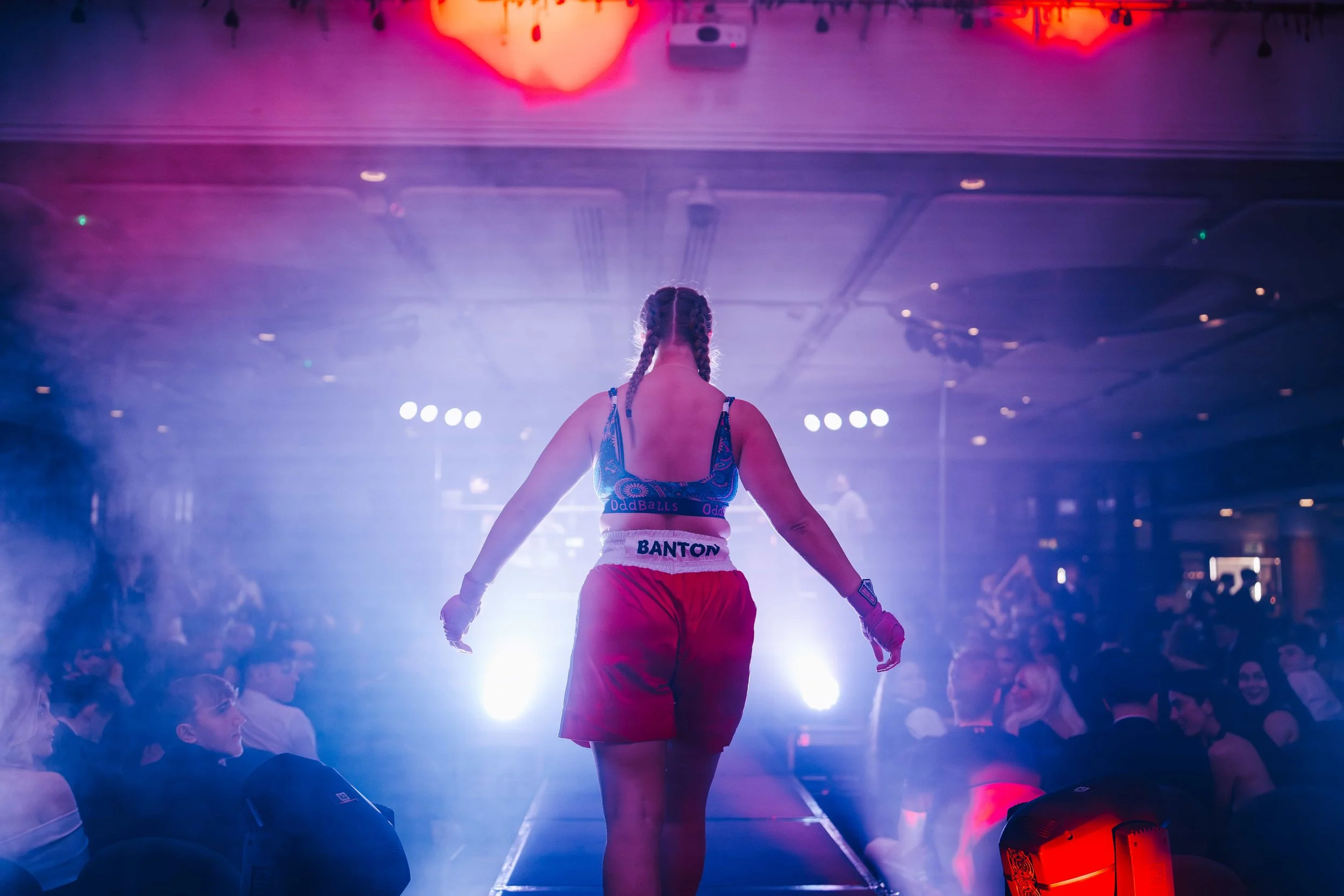 Female boxer with braided hair walking out to the ring on a runway with a crowd watching in the background, lit with blue and purple stage lighting.