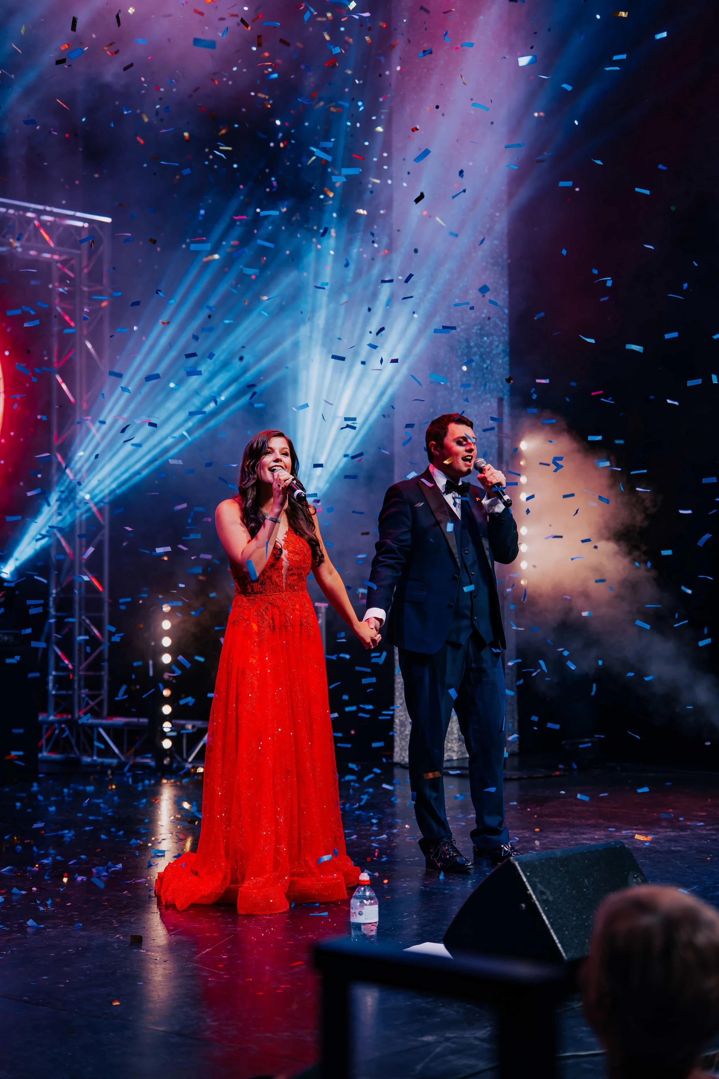 A man and woman dressed in formal attire holding hands on a stage with bright lights and confetti falling around them.