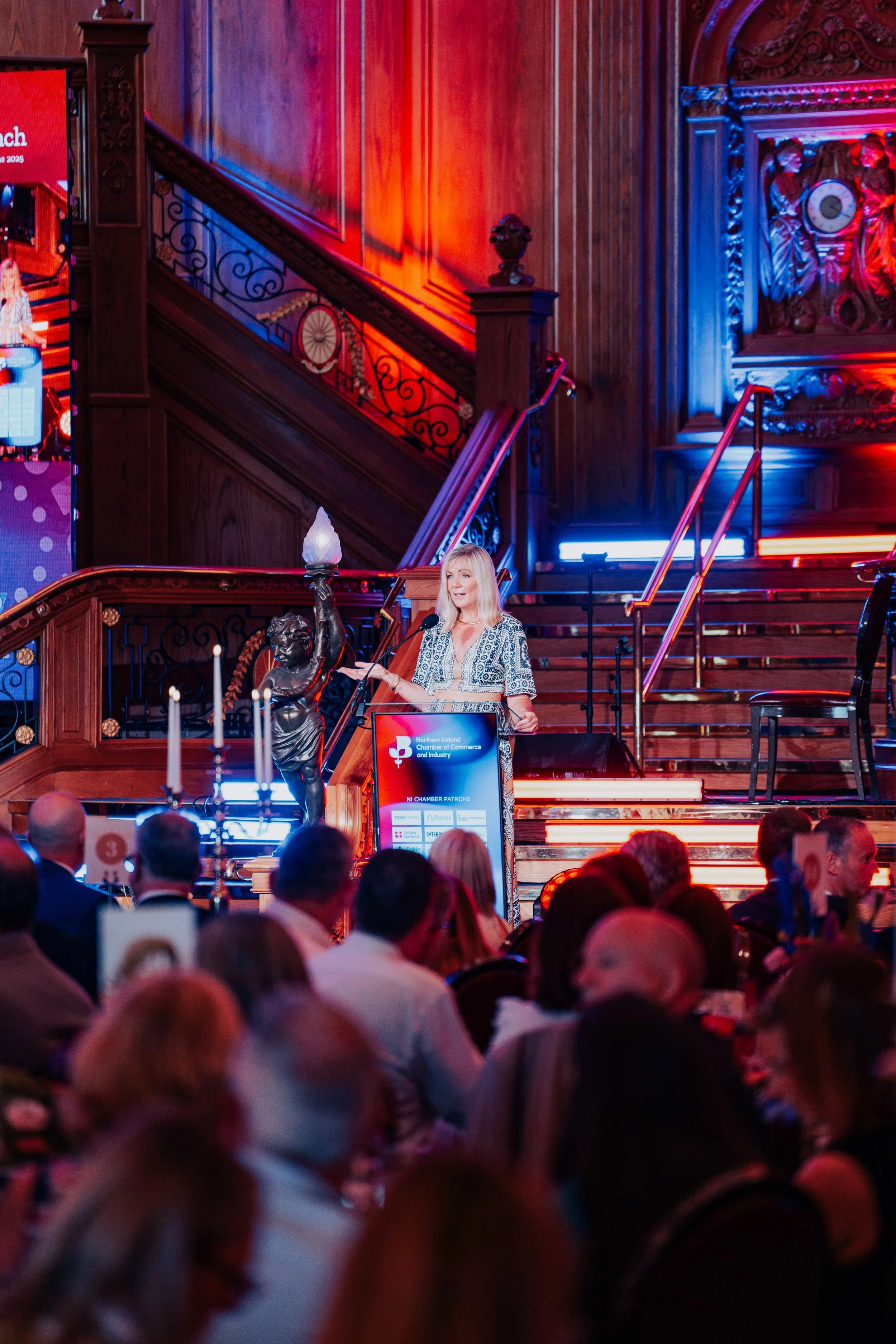 A woman giving a speech at a formal event on a stage in a grand, wood-paneled hall with ornate decorations. The stage has a podium, large sculpture of a cherub holding a flame, and colorful lighting. Audience members are seated at tables facing the stage.