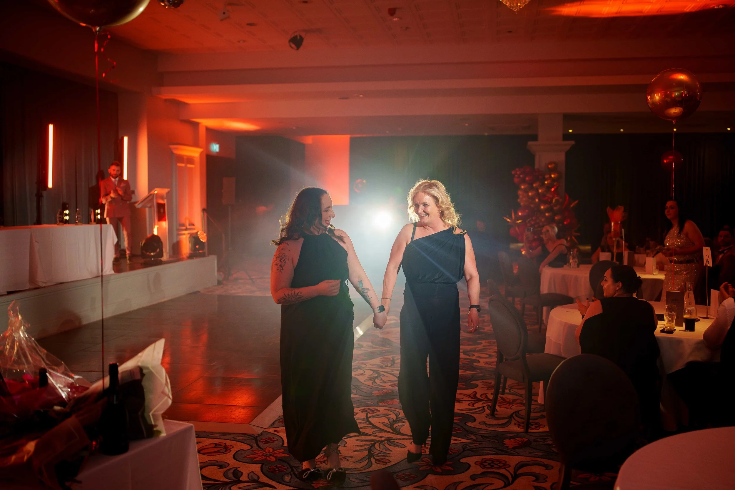 Two women dancing hand-in-hand at an indoor party or event with tables, seated guests, a stage, lighting effects, and decorative balloons in the background.