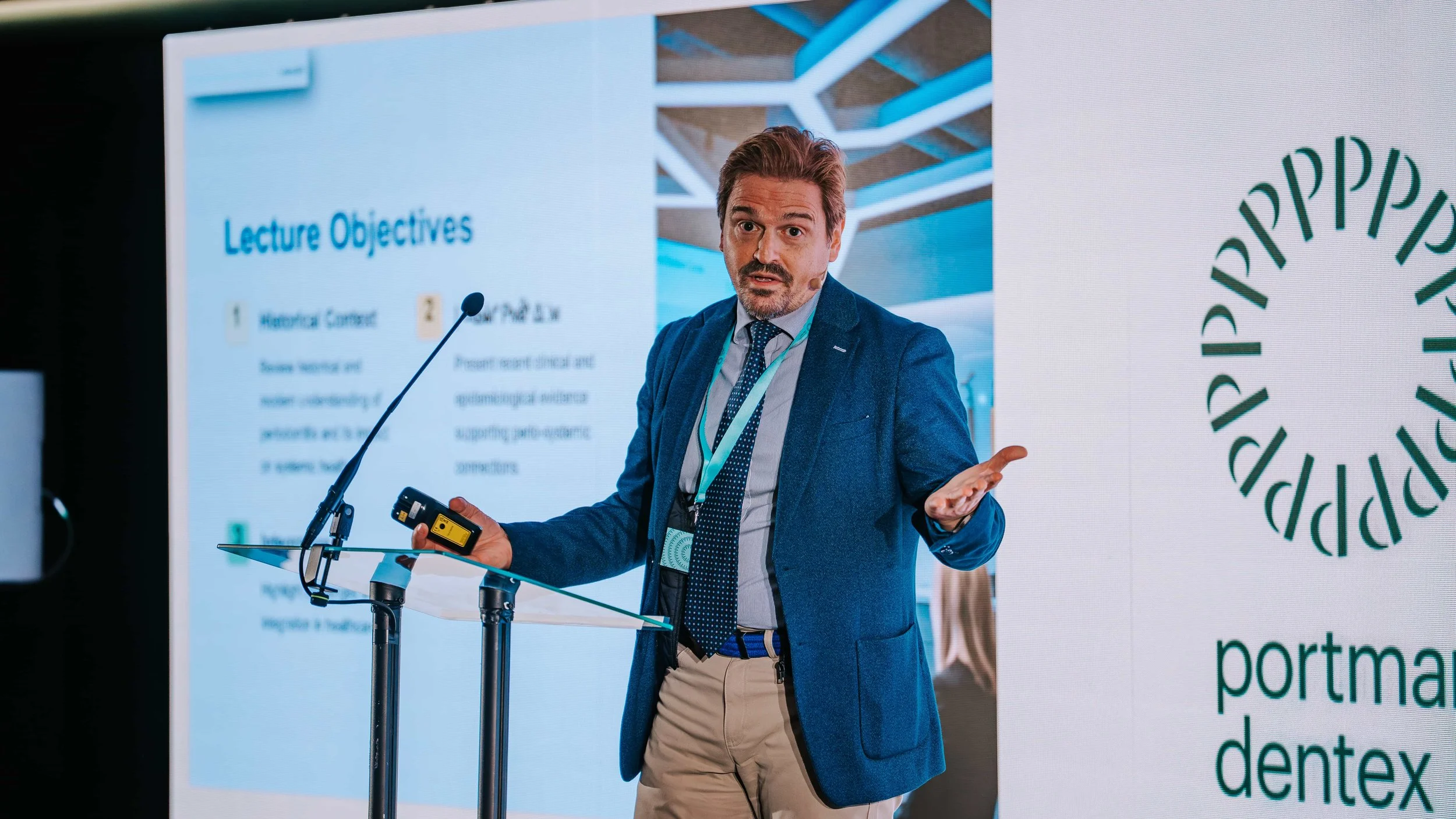 Man in blue blazer and beige pants giving a presentation at a conference, standing at a transparent lectern with a microphone, digital clicker in hand, gesturing with his left hand, with a large presentation slide behind him displaying the words 'Lec