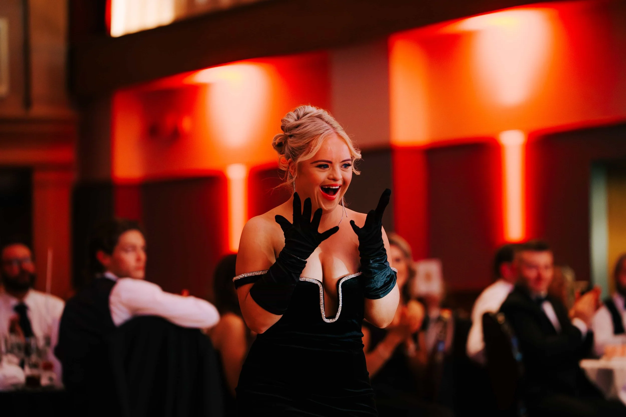 A woman with blond hair in an updo wearing a black dress and long black gloves, smiling and reacting with excitement at an event with an audience in a dimly lit room with red and orange lighting.