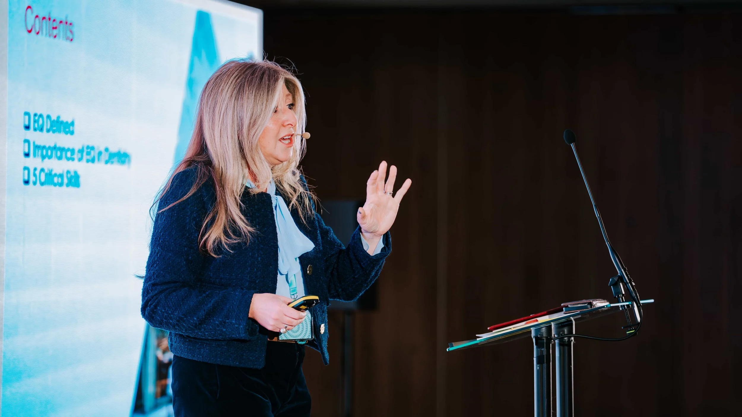 A woman with long blonde hair giving a presentation on stage. She is wearing a dark blue blazer and holding a remote control. There is a microphone attached to her face, and a large screen displaying text behind her. The background is dark.