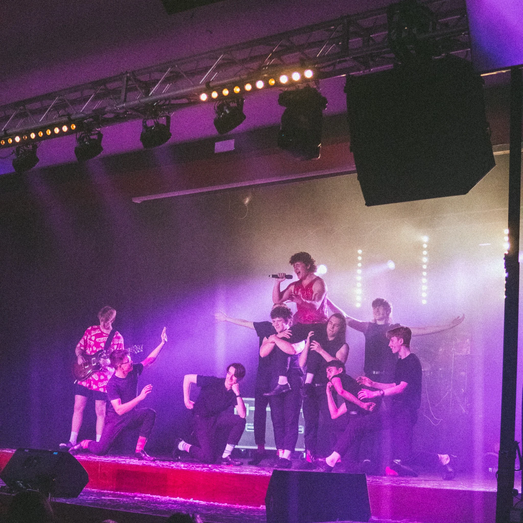A group of performers on a stage forming a human pyramid, with one person standing on top holding a microphone, surrounded by others in various poses; stage lights and curtains in the background.