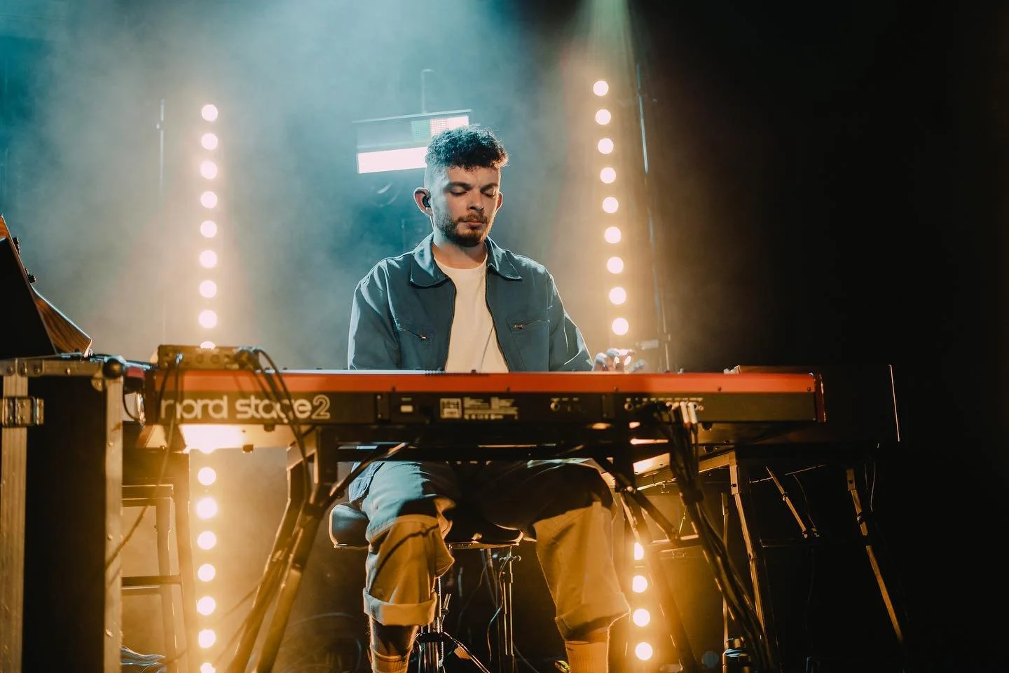 A male musician playing a digital keyboard on stage with stage lights and fog.