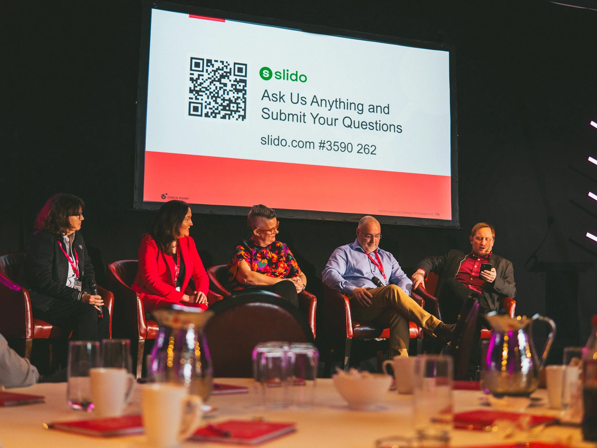 Panel of five individuals sitting on chairs on a stage at a conference, with a large screen behind them displaying a QR code and information about Slido.