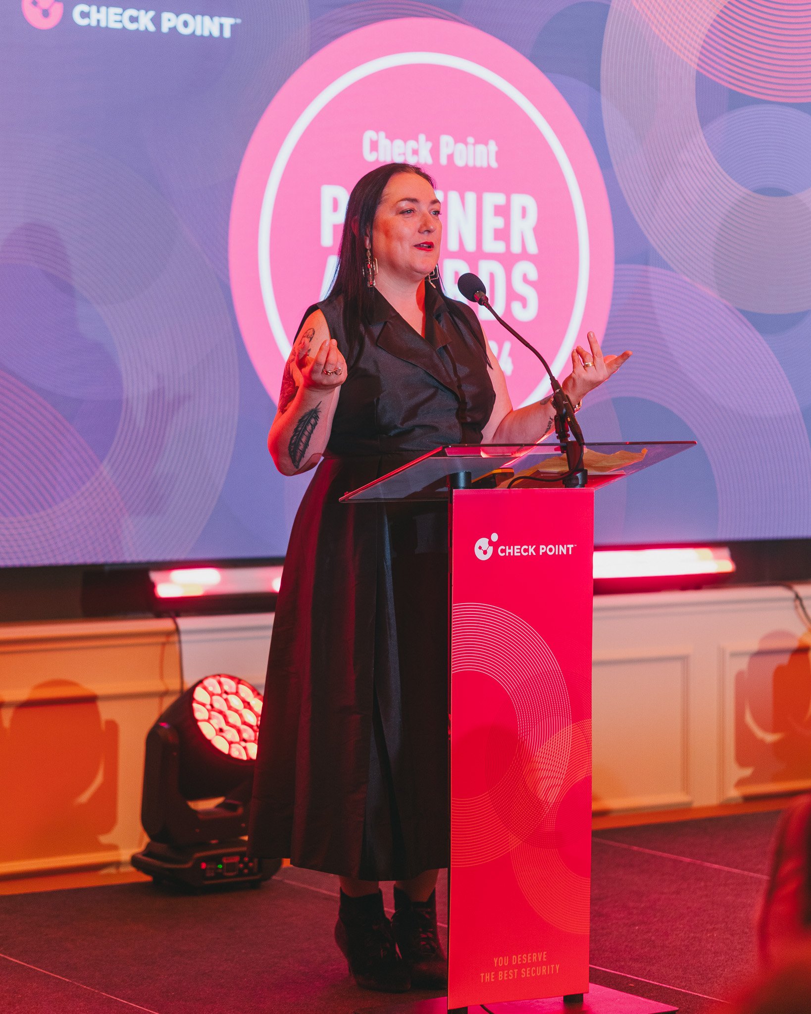 A woman with dark hair and tattoos on her arms giving a speech at a podium with a Check Point logo, standing in front of a large pink and purple screen that says "Check Point Partner Awards."
