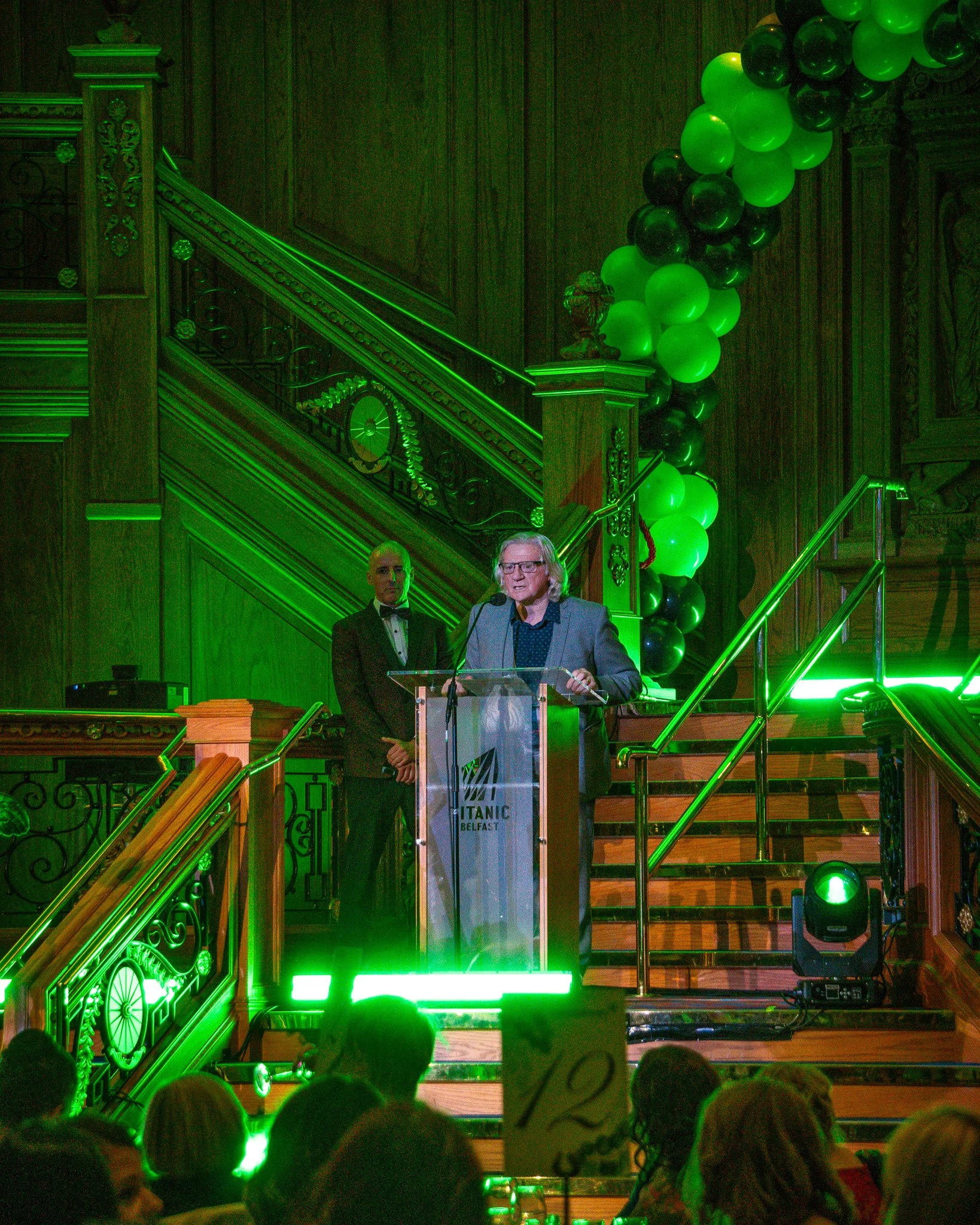 A man in glasses and a gray suit speaks at a podium at an event with the Titanic Belfast logo. A man in a tuxedo stands behind him. The stage is decorated with green balloons and green lighting, with a wooden staircase and ornate wood paneling in the
