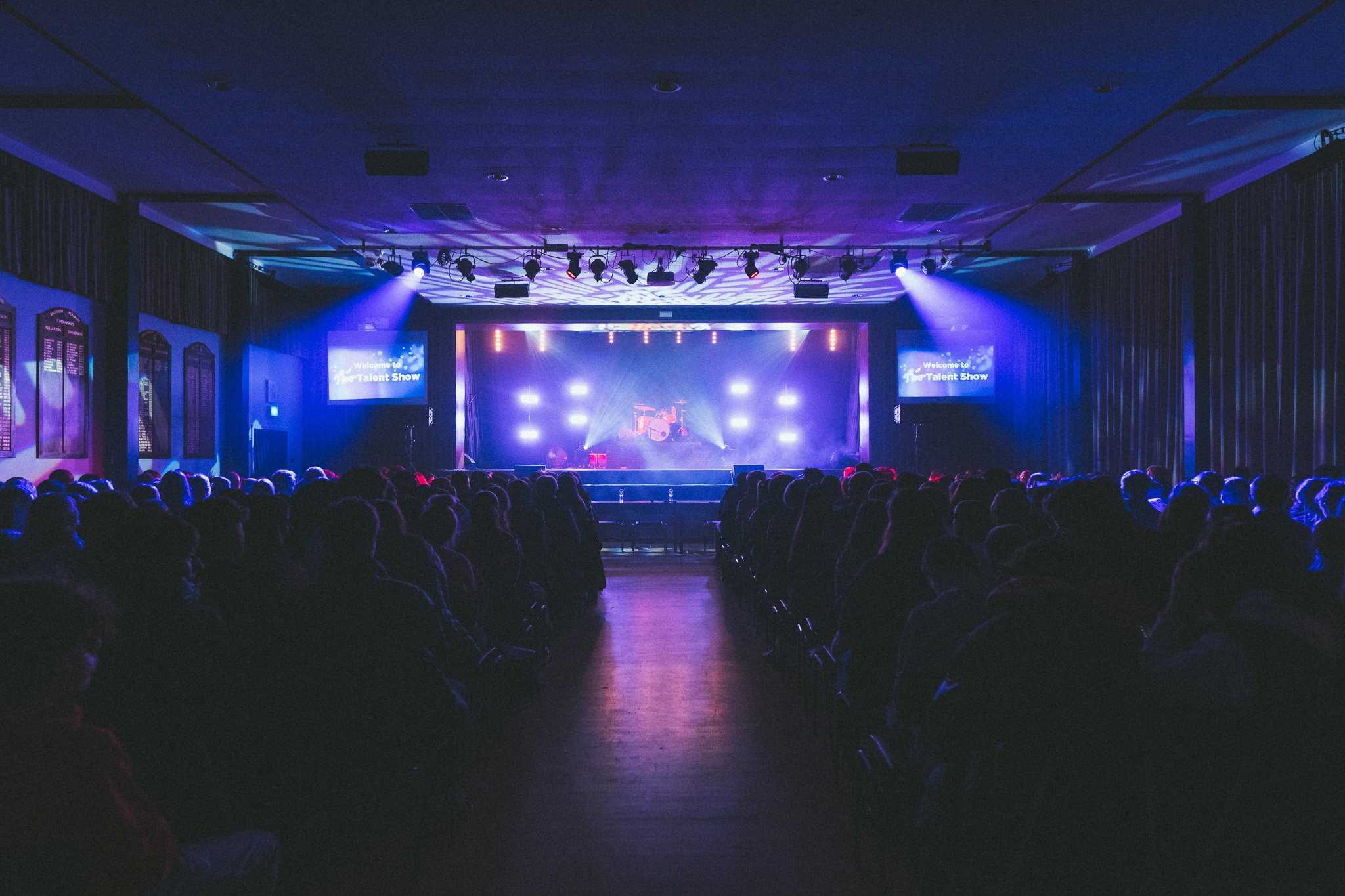 A dark theater with an audience watching a stage with purple and blue lights, and a drum set at the center. Two screens on each side display 'Welcome to the Talent Show'.