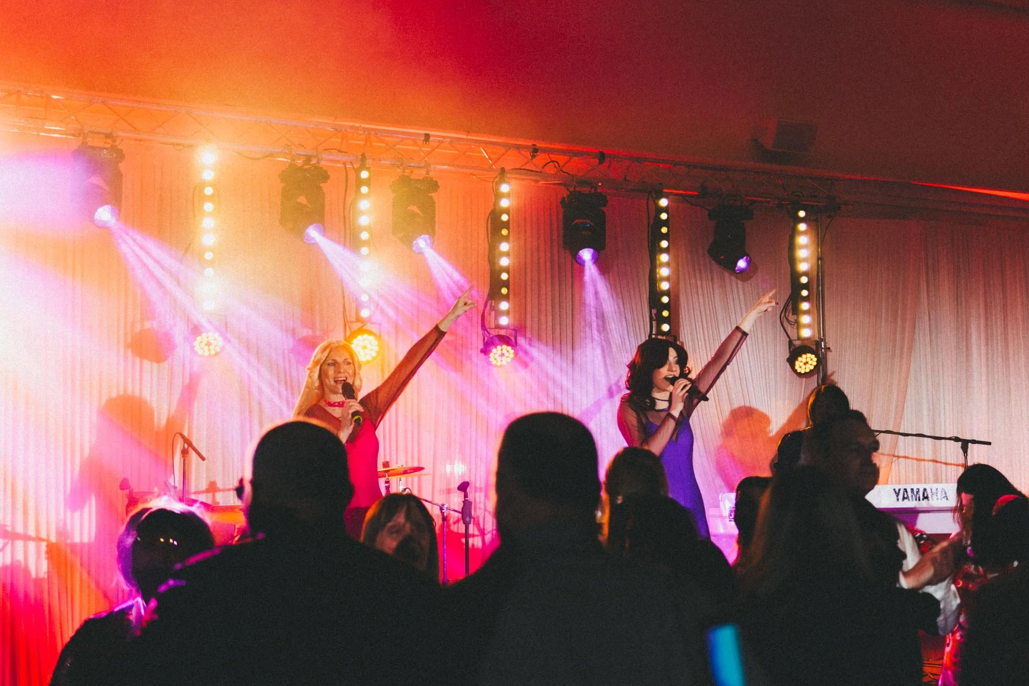 Two women singing on stage with colorful lights, one with long blonde hair wearing pink, and the other with dark hair wearing purple, in front of an audience.