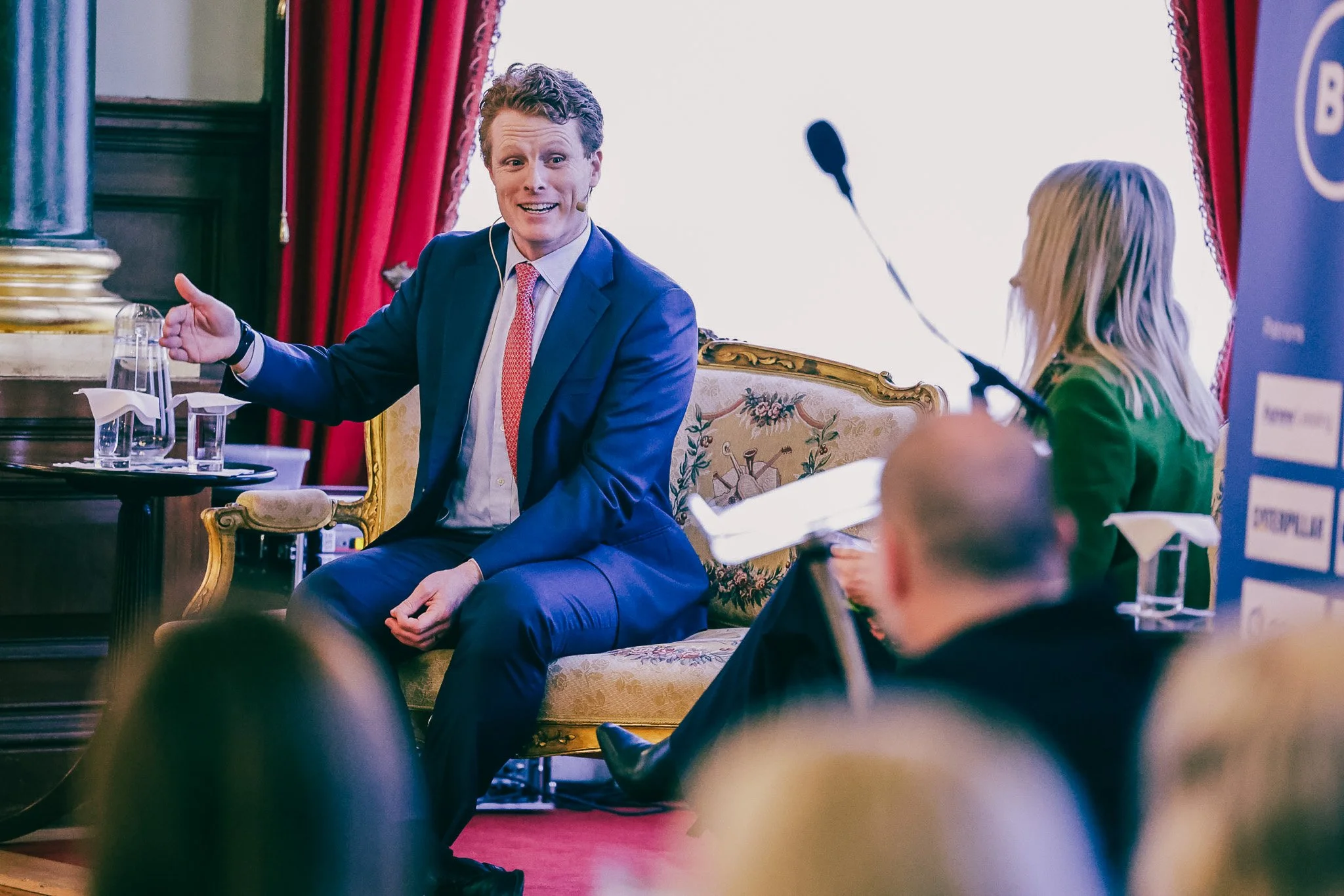 A man in a blue suit with a pink tie speaking at a conference, sitting on an ornate chair, with a woman nearby taking notes and a microphone in her hand, in a room with red curtains.