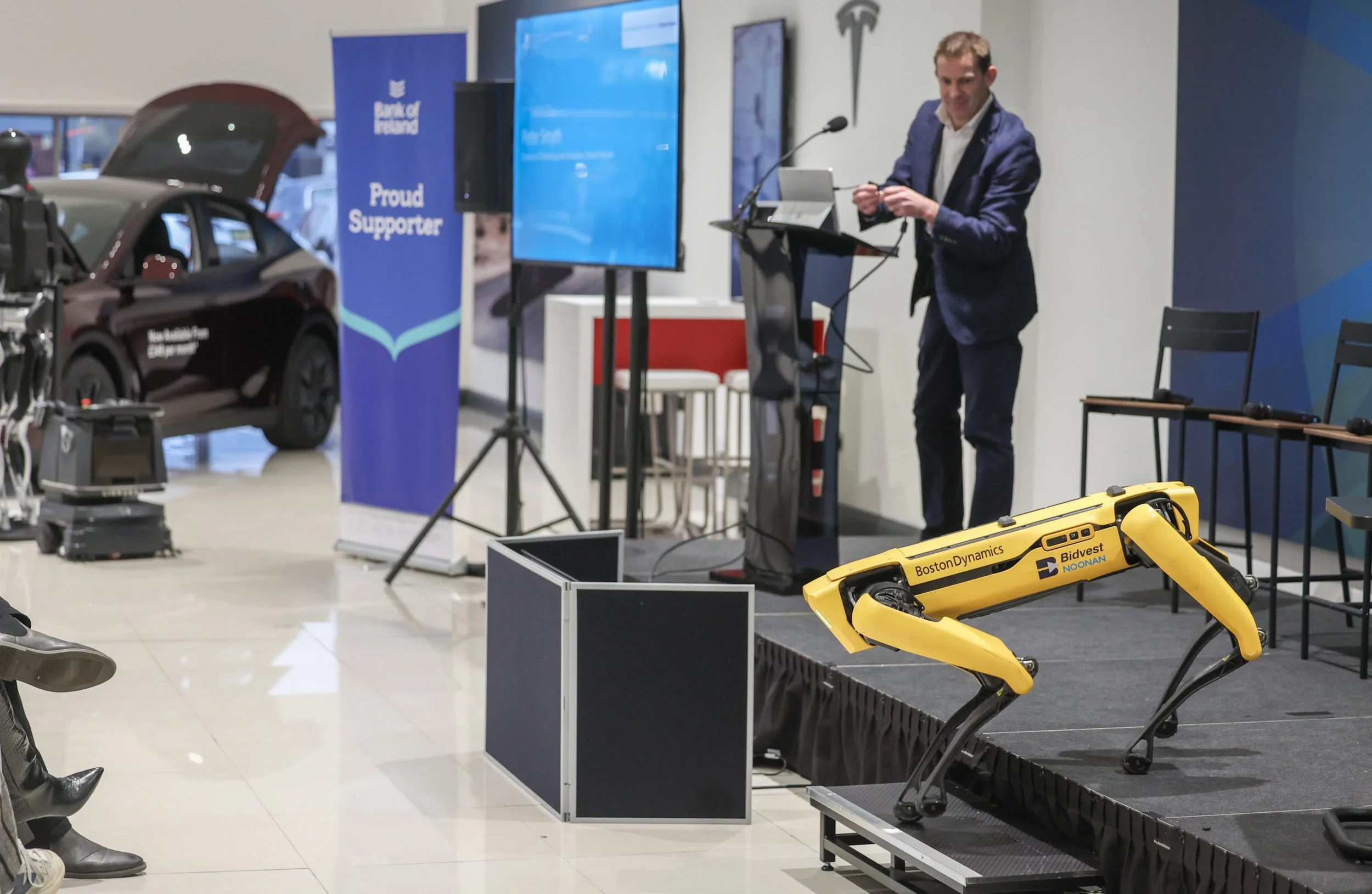 A man in a suit stands on a stage with a laptop, speaking at a presentation event. There is a yellow robotic dog on the stage, and a car with its door open is visible behind the stage. A blue banner with the logo 'Bank of Ireland' and text 'Proud Sup