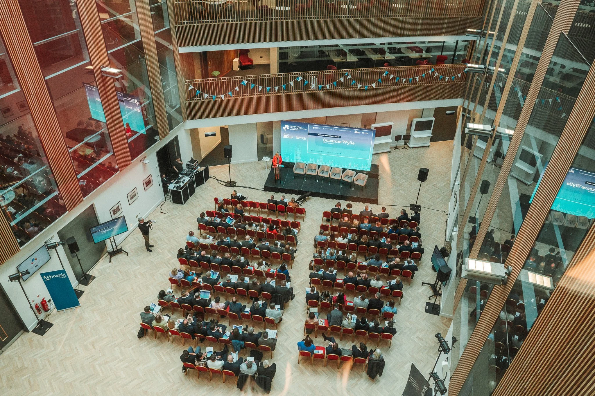 An auditorium with a panel of speakers at the front and an audience seated facing the stage. There are multiple screens and cameras set up around the room.