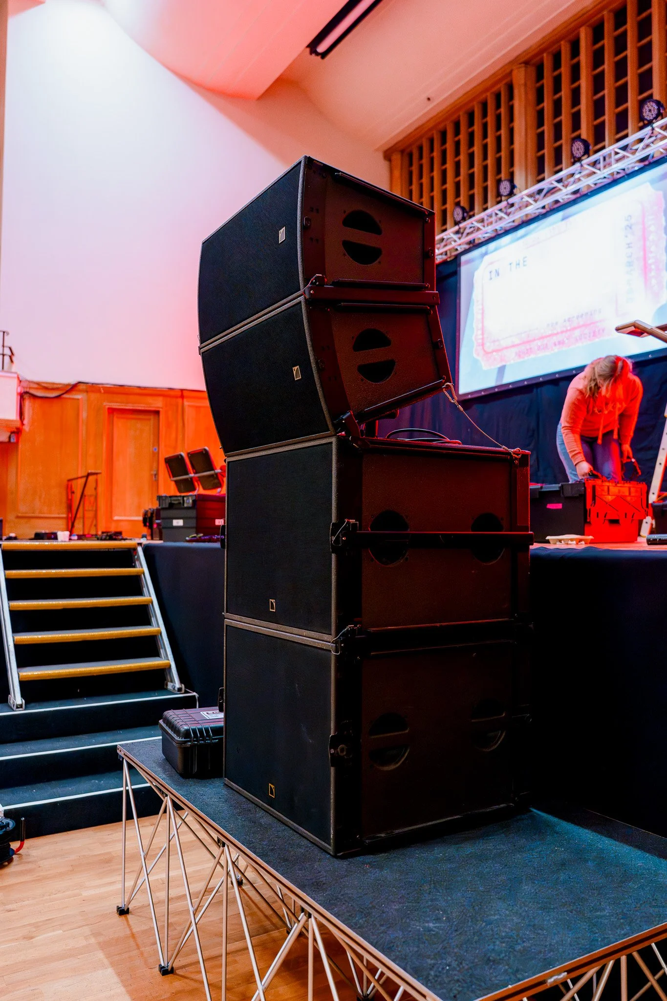 A stack of black speakers on a platform at an indoor event with a large screen and a woman setting up in the background.