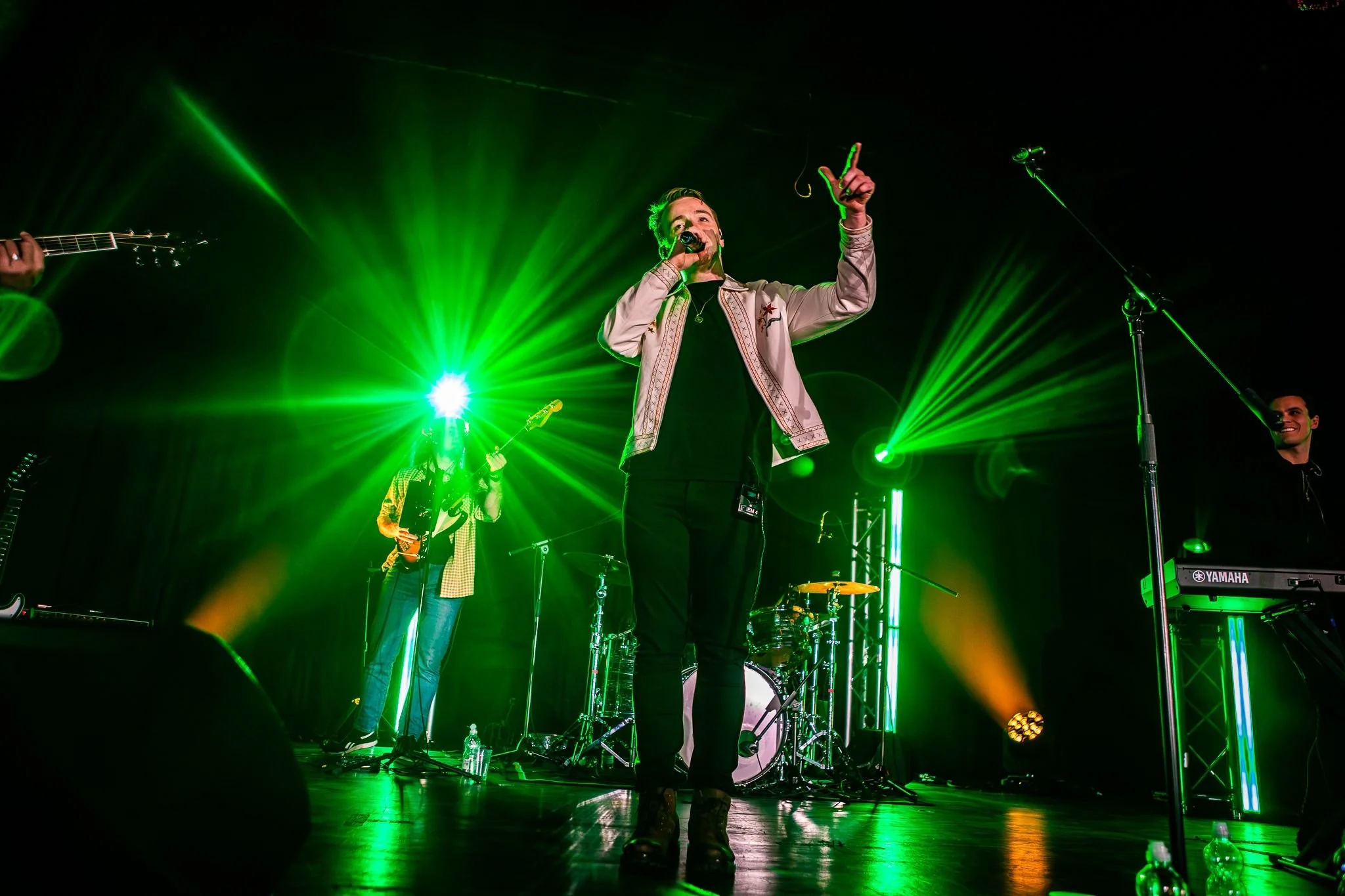 Performer singing on stage with a microphone, surrounded by green laser lights, with band members playing instruments in the background.