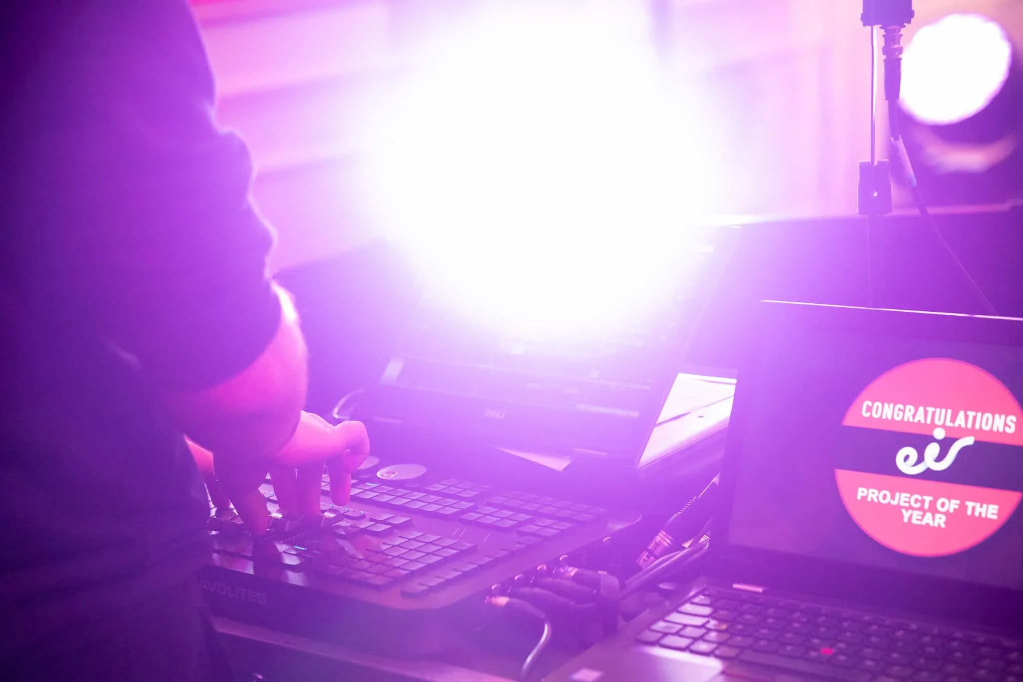 Person working on a laptop with a bright purple light behind them, indicating a celebratory event, with a pink circle in the bottom right that says 'Congratulations' and 'Project of the Year'.