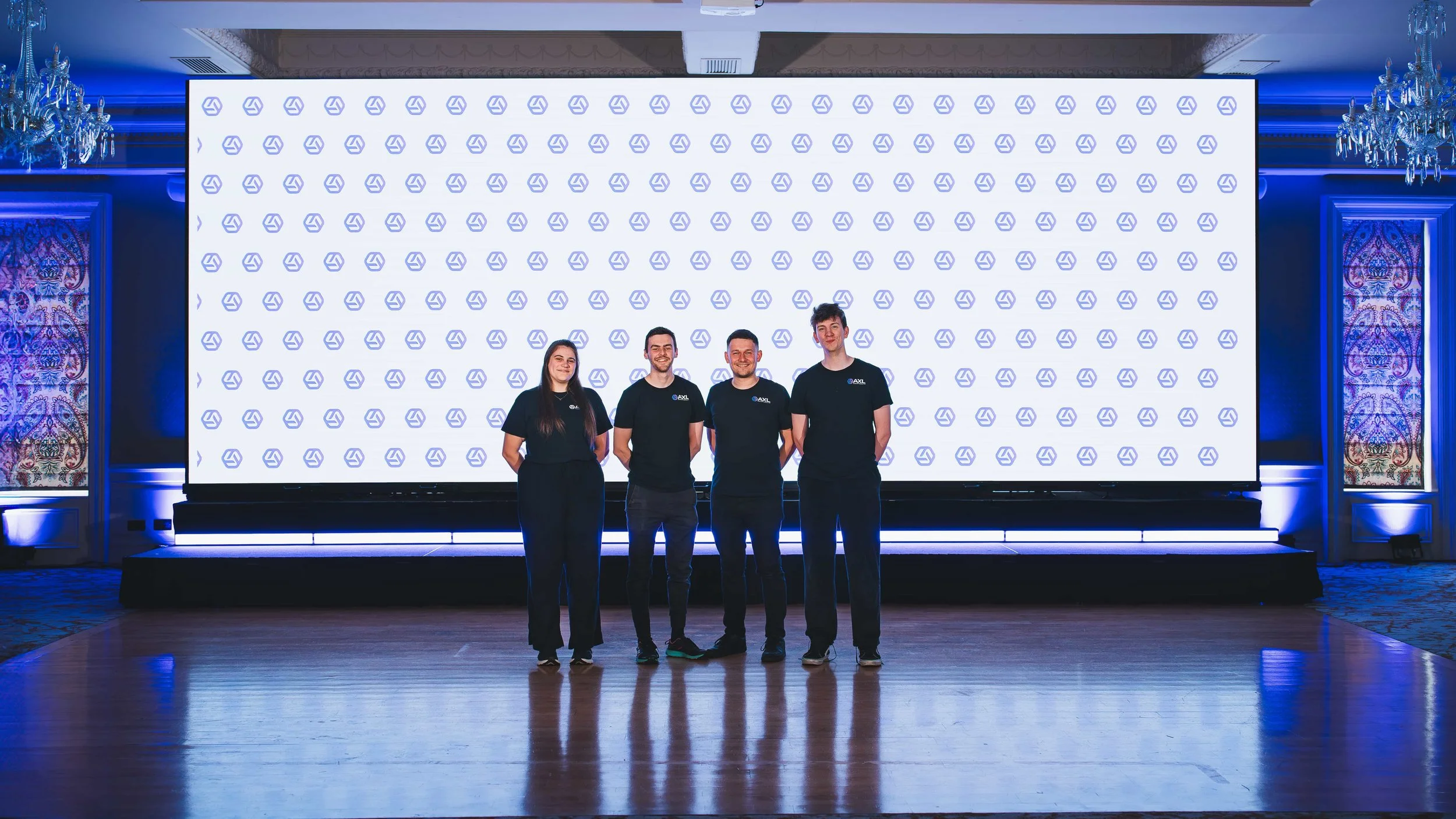 Four people standing in front of a large screen with a repeated logo pattern, in a decorated conference or event room with chandeliers and ornate curtains.