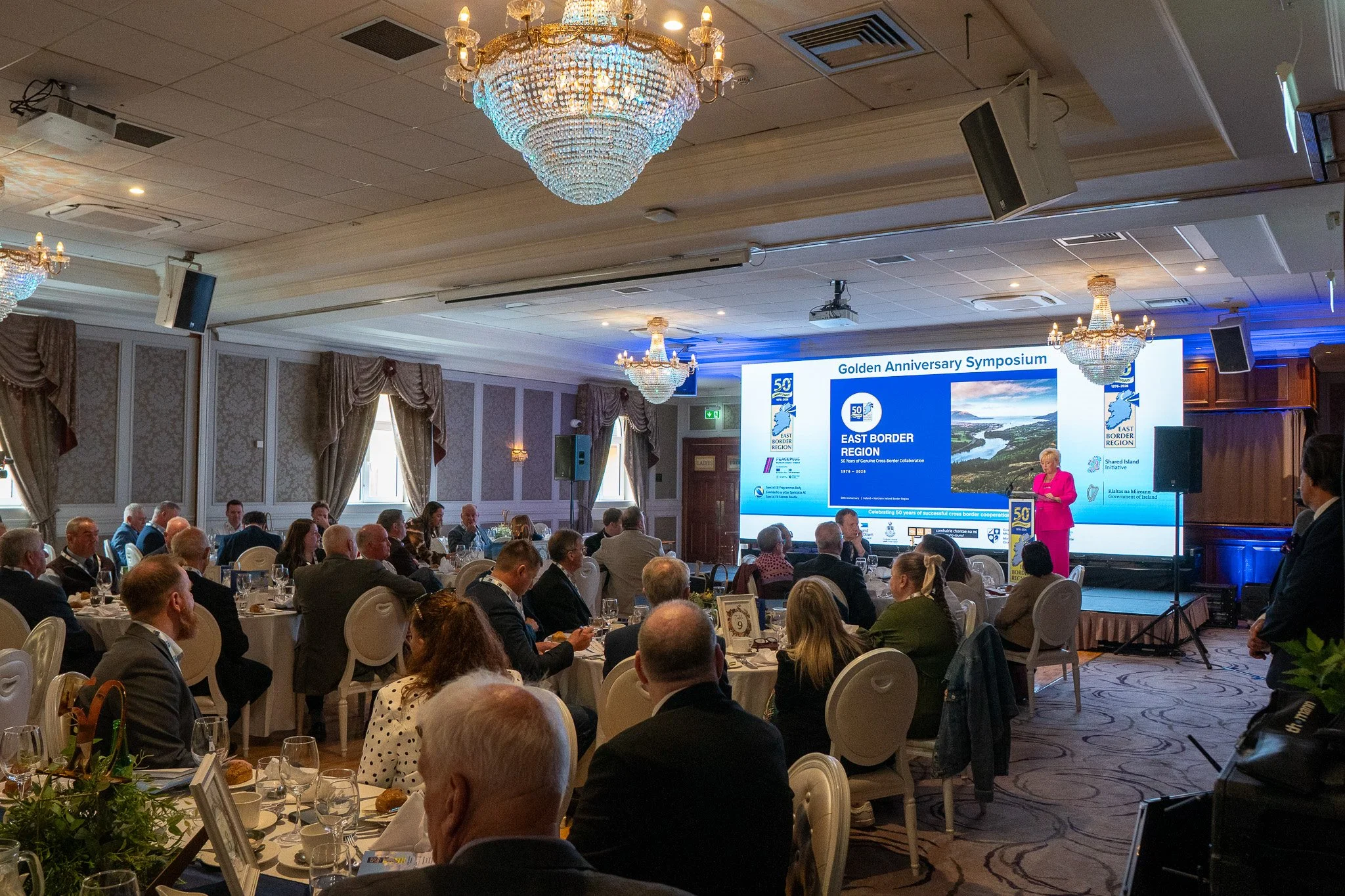 Golden Anniversary Symposium at Canal Court Hotel Newry, speaker at lectern before a 6m LED video wall and round lunch tables.