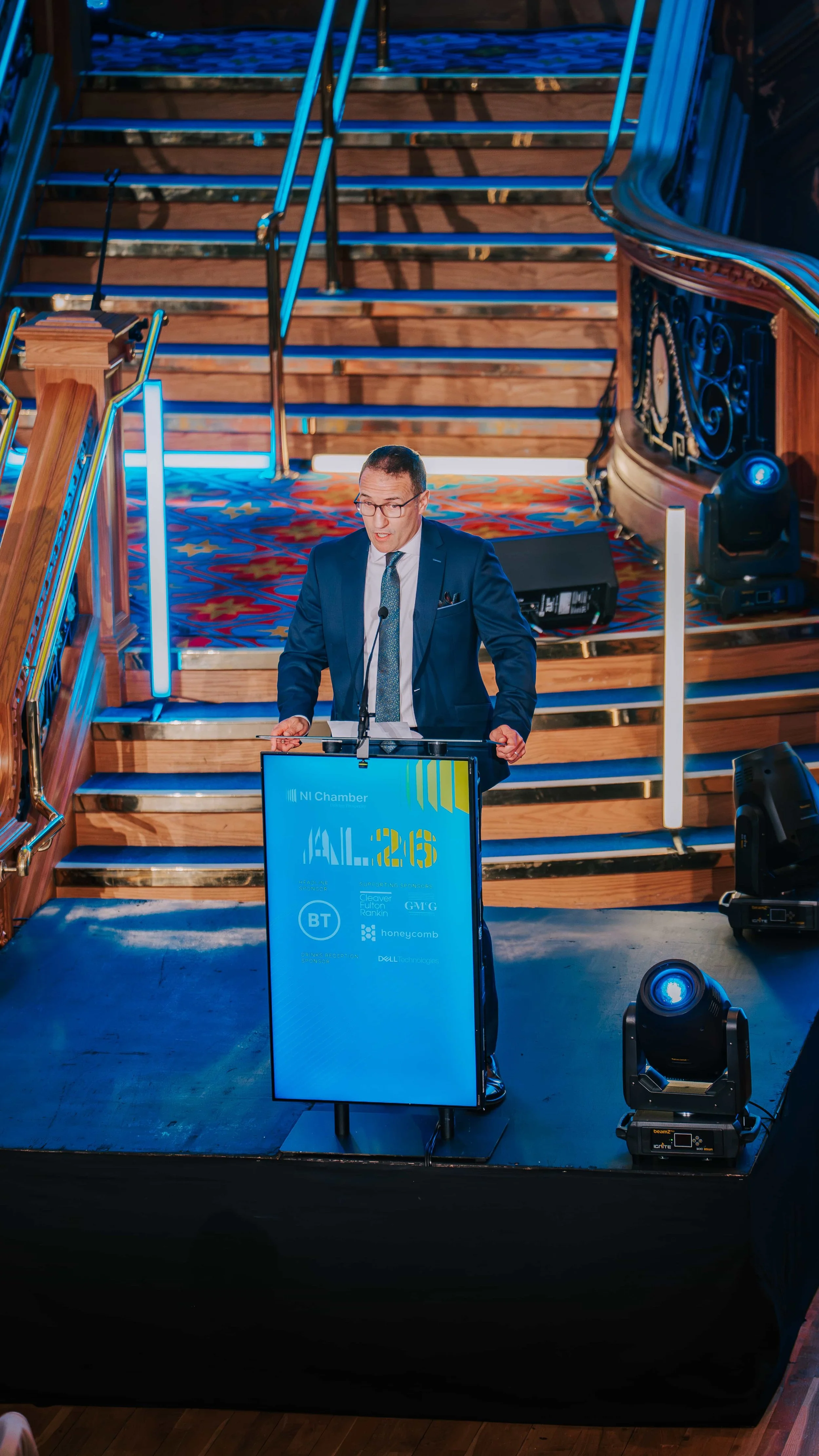 Man in a suit speaking at a podium during a conference, with a staircase in the background, on a stage with lighting equipment around.