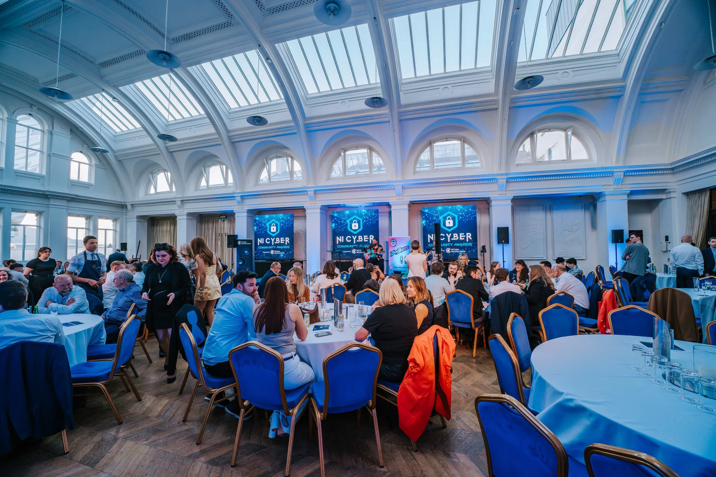 An indoor event at a grand hall with a high glass ceiling, where people are seated at round tables, some facing a stage with LED screens displaying 'NICYBER Community Awards,' and a musician performing.