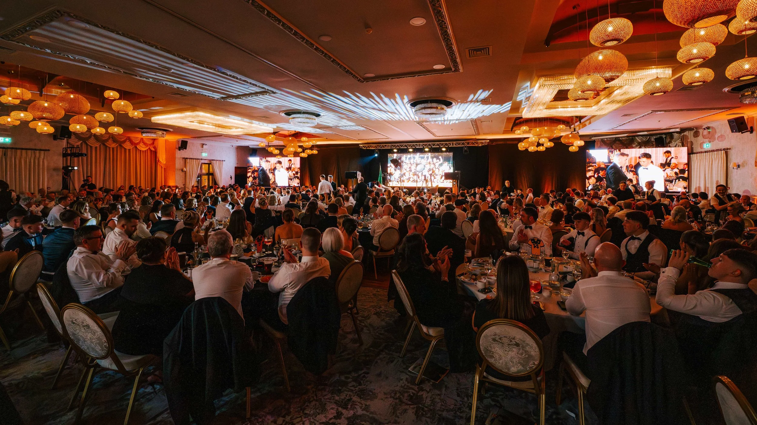A large banquet hall filled with people dressed in formal attire, seated at round tables, attending a formal event or celebration. There is a stage with large screens and lighting effects.
