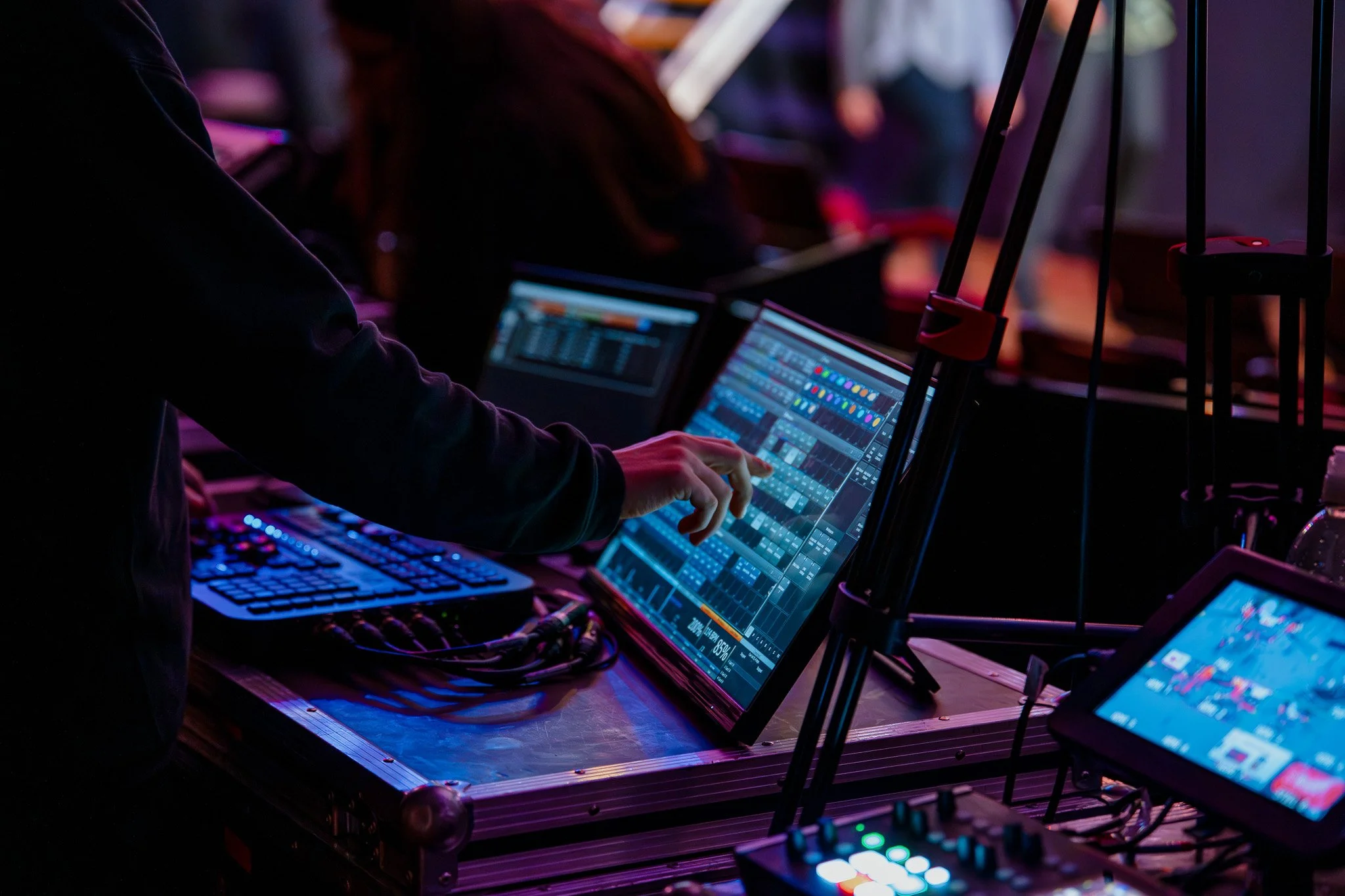 A DJ mixes music at a nightclub with colorful lighting, using a touchscreen controller, laptop, and other electronic equipment on a table.