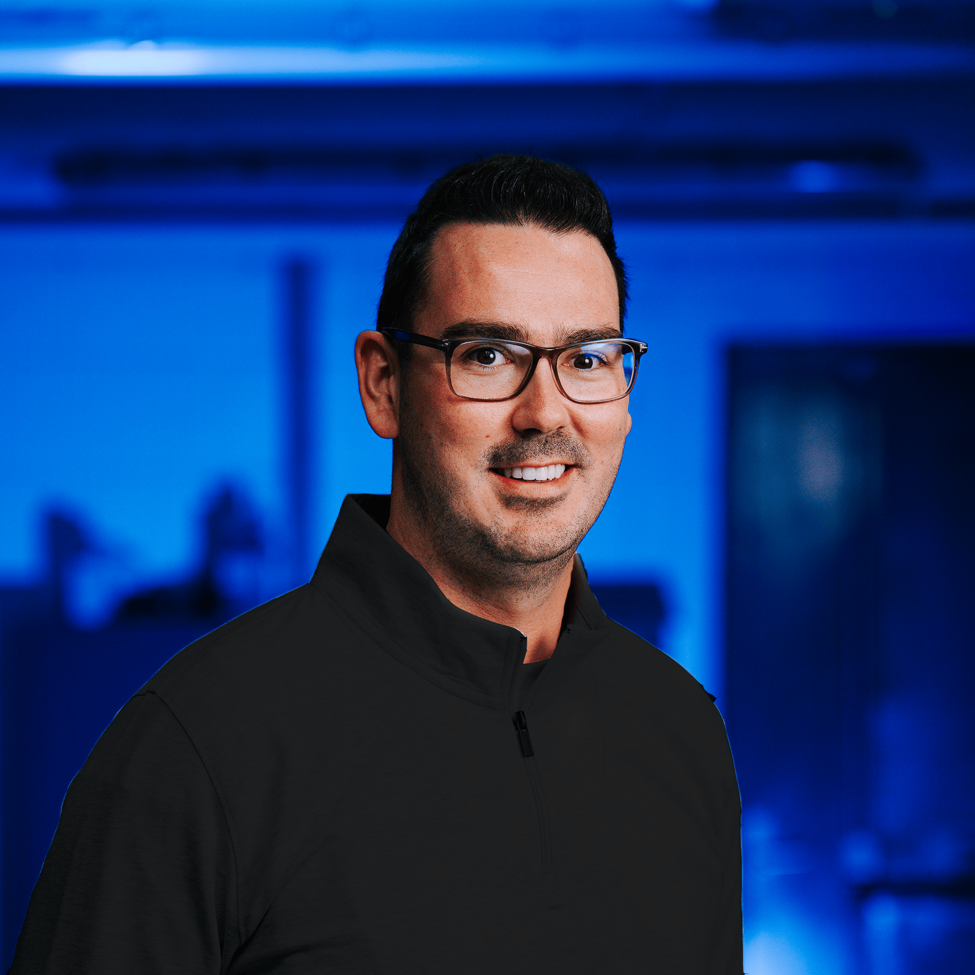 A man wearing glasses and a black zip-up jacket smiling in a setting with blue lighting.