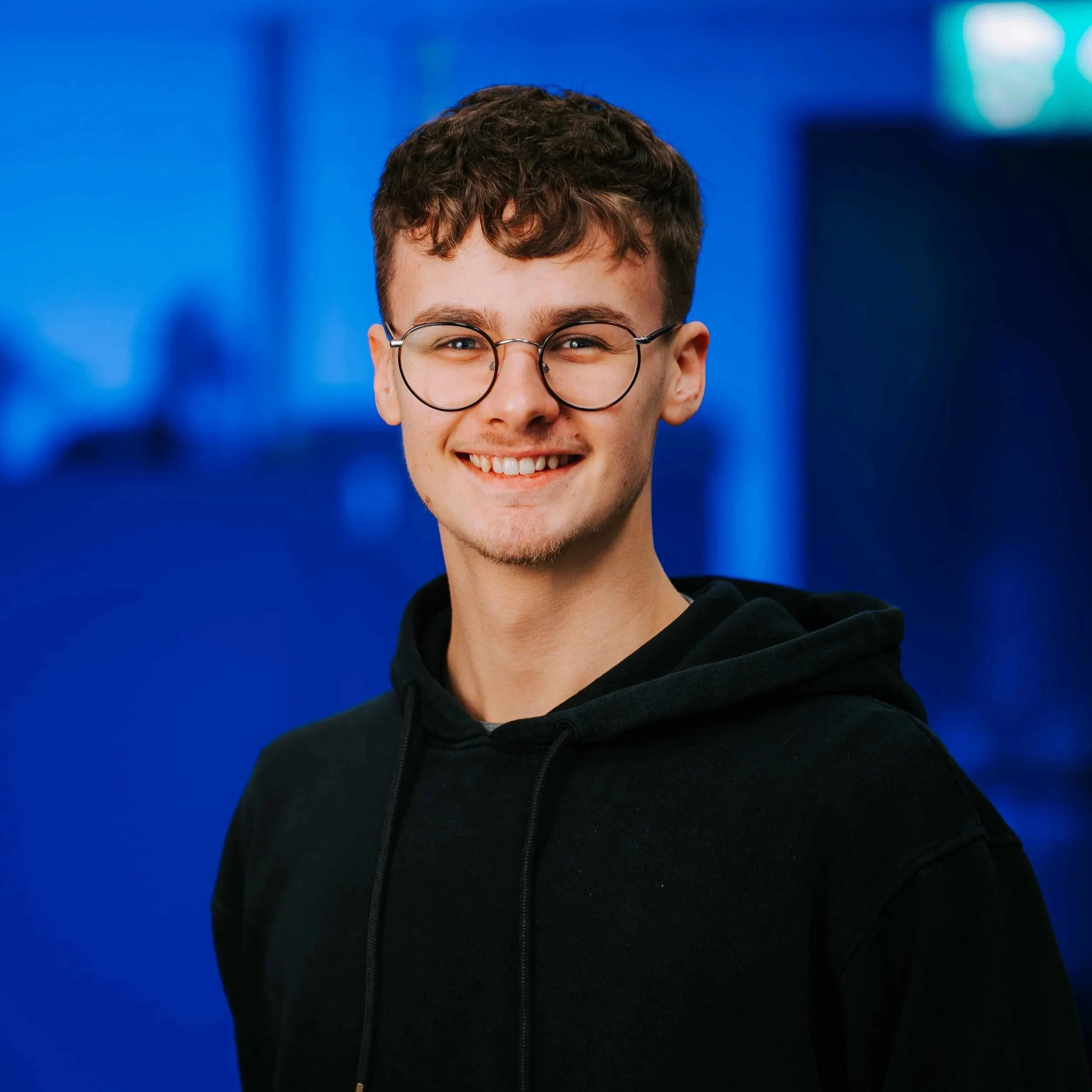 Smiling young man with glasses wearing a black hoodie against a blue background.