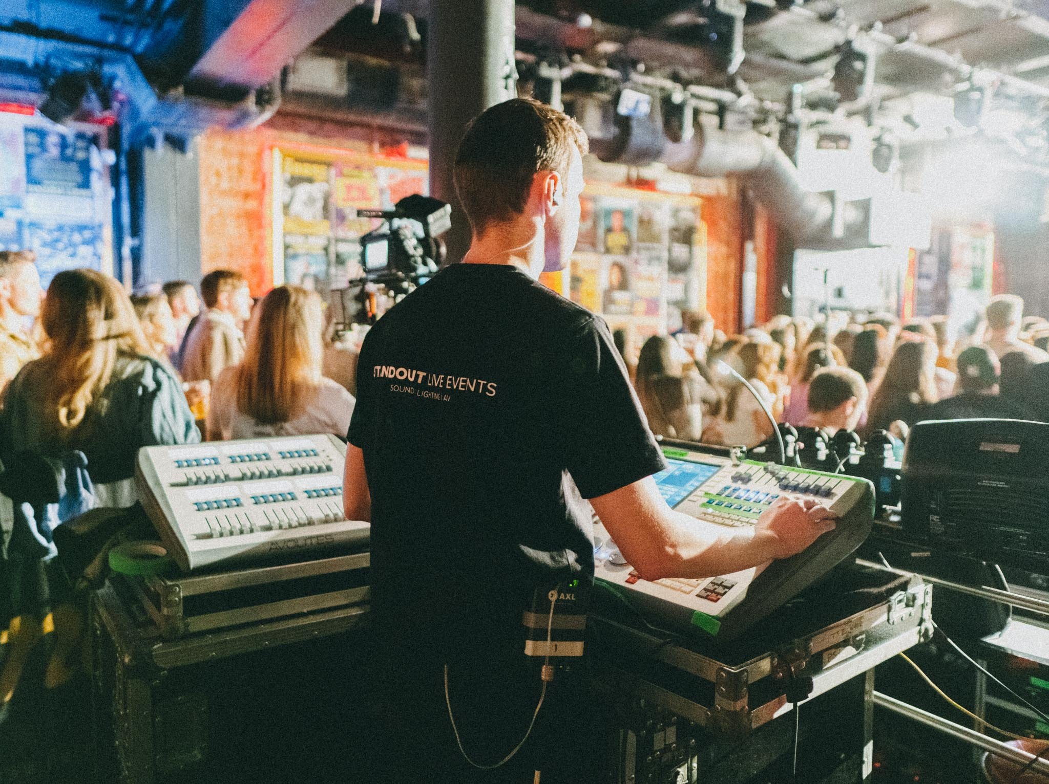 A DJ is playing music at a live event with a crowd of people watching in an indoor venue. The DJ is standing behind a mixing console, wearing a black t-shirt with the words 'STANDOUT LIVE EVENTS' on it.