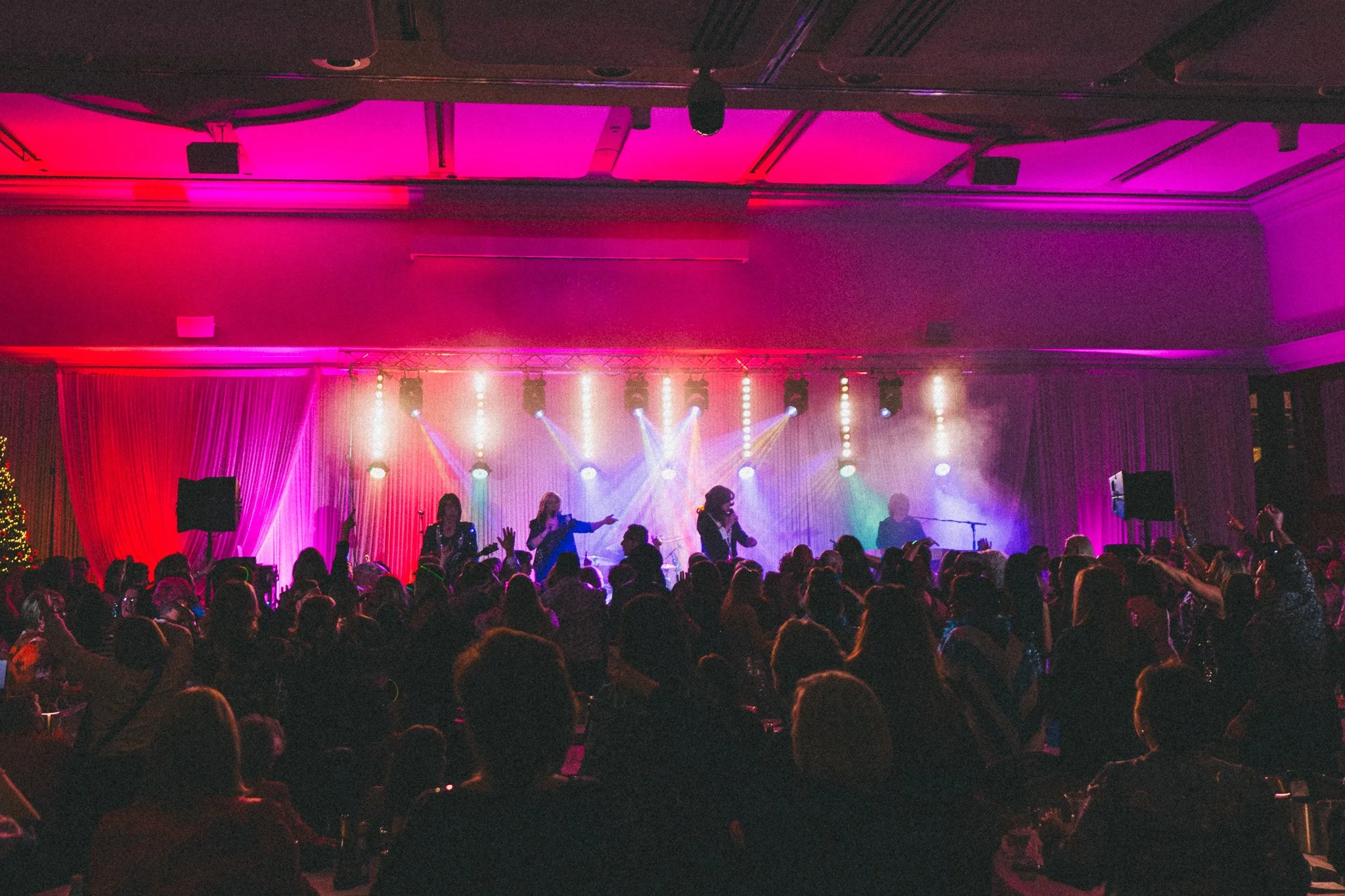 Concert scene with a stage, colorful lights, and performers singing; audience watching and recording with phones in a large indoor venue.