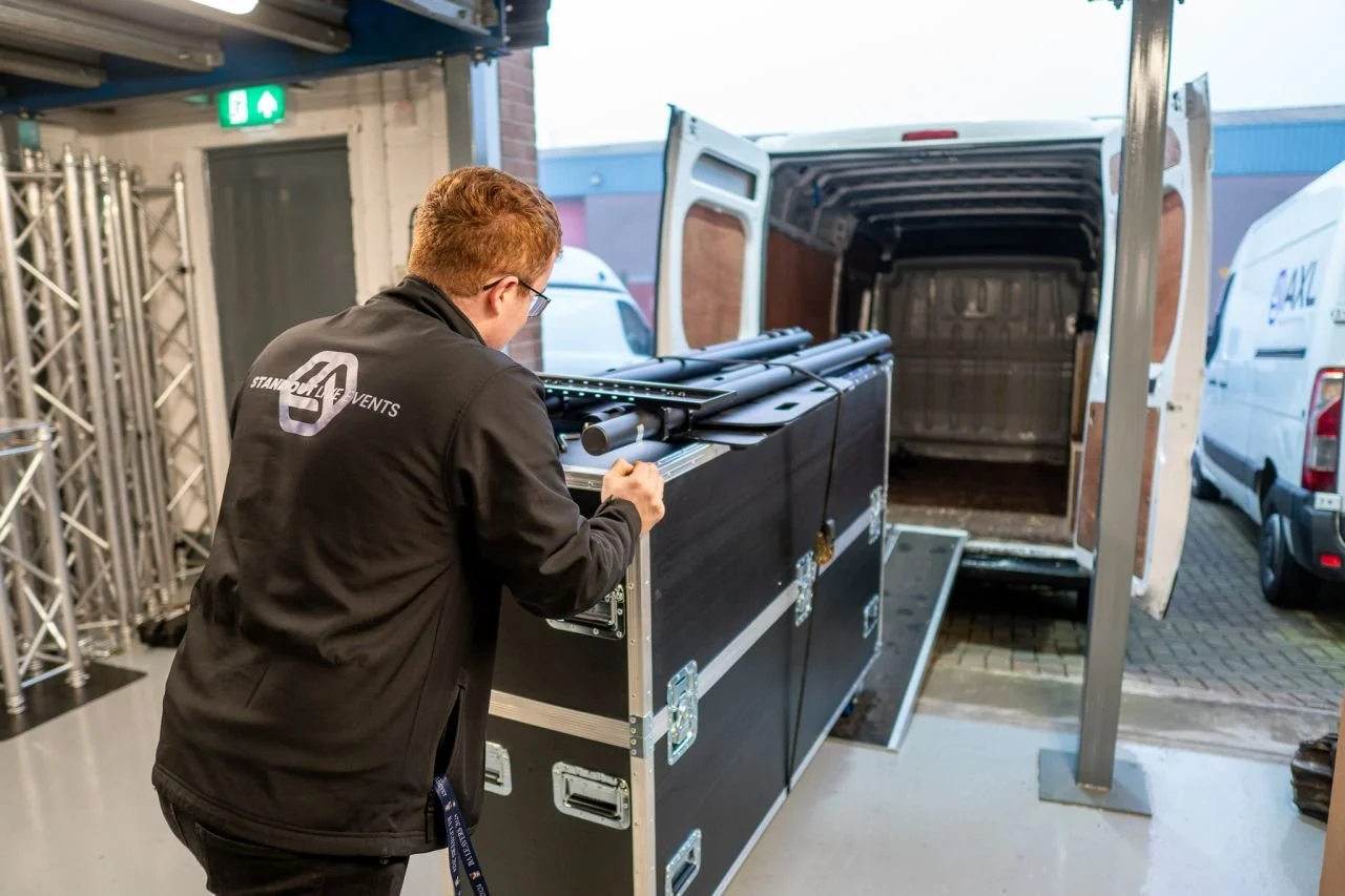 A man with red hair and glasses wearing a black jacket with the logo 'STAND OUT EVENTS' is working on a large black equipment case near an open van. The van's back doors are open, revealing an empty cargo space. There are metal trusses on the left side and a green emergency exit sign above a door in the background.