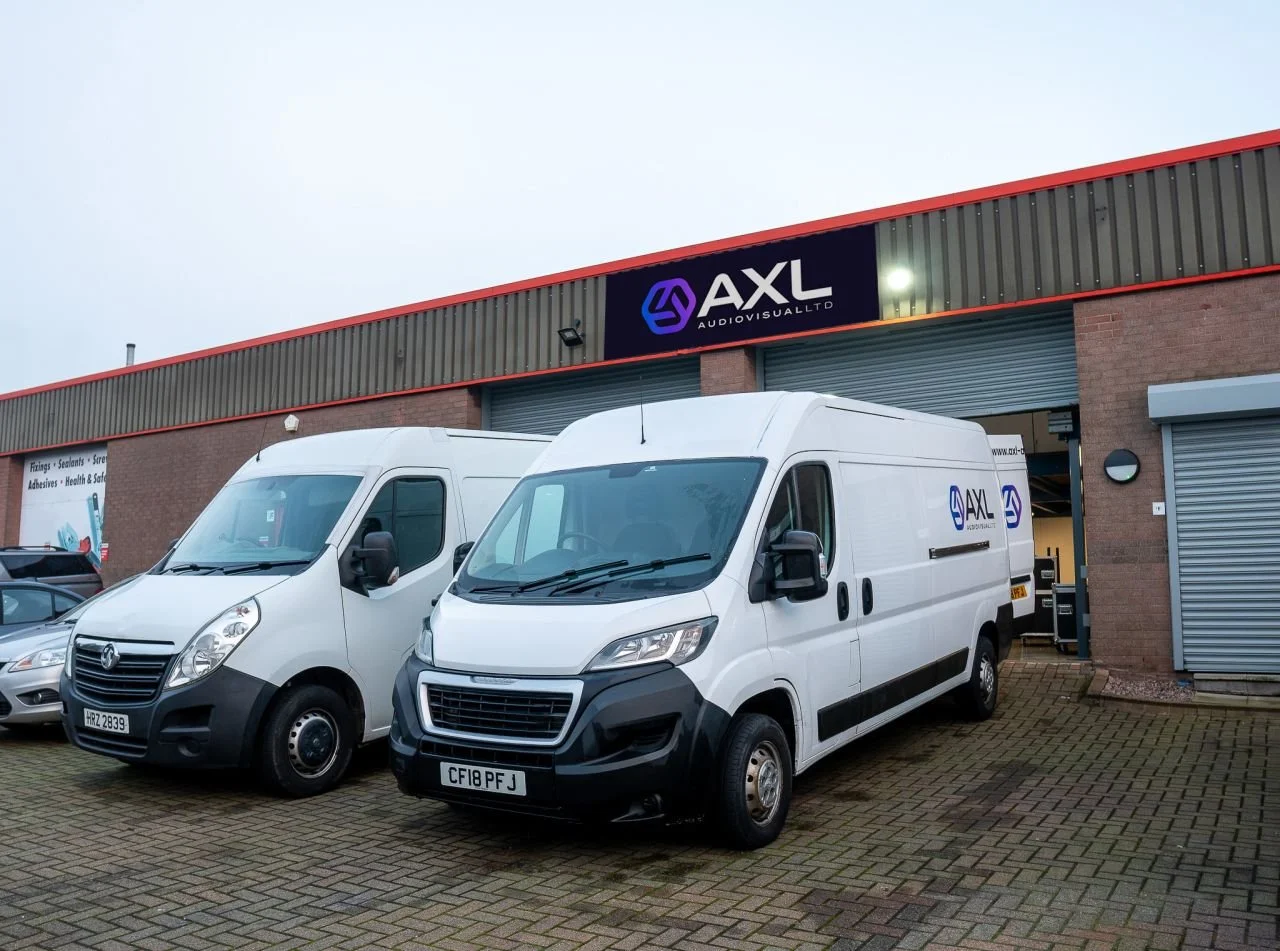 Two white commercial vans parked in front of a building with a sign for AXL Audiovisual Ltd. The building has a brick exterior with a red trim at the top and gray roller doors.