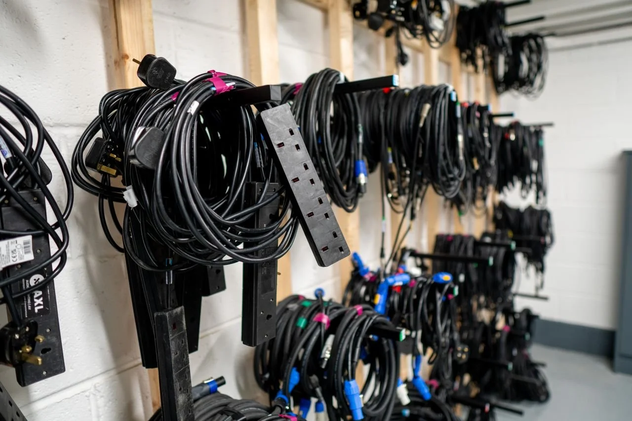 A wall with multiple hooks holding neatly coiled black cables, some with colorful connectors, in what appears to be a workshop or storage room.