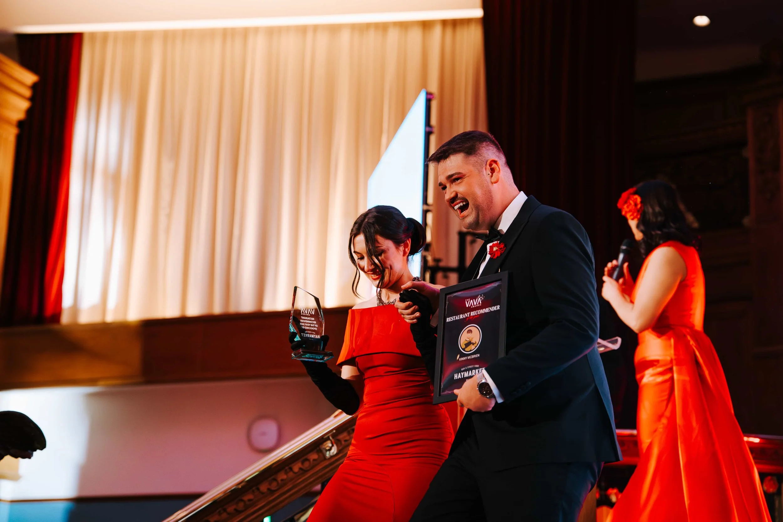 Three people at an awards ceremony. A man in a tuxedo and two women in red dresses are smiling and holding awards. One woman is looking down, while the man is looking ahead. The woman in the background has a microphone and is dressed in a red dress with a flower in her hair.