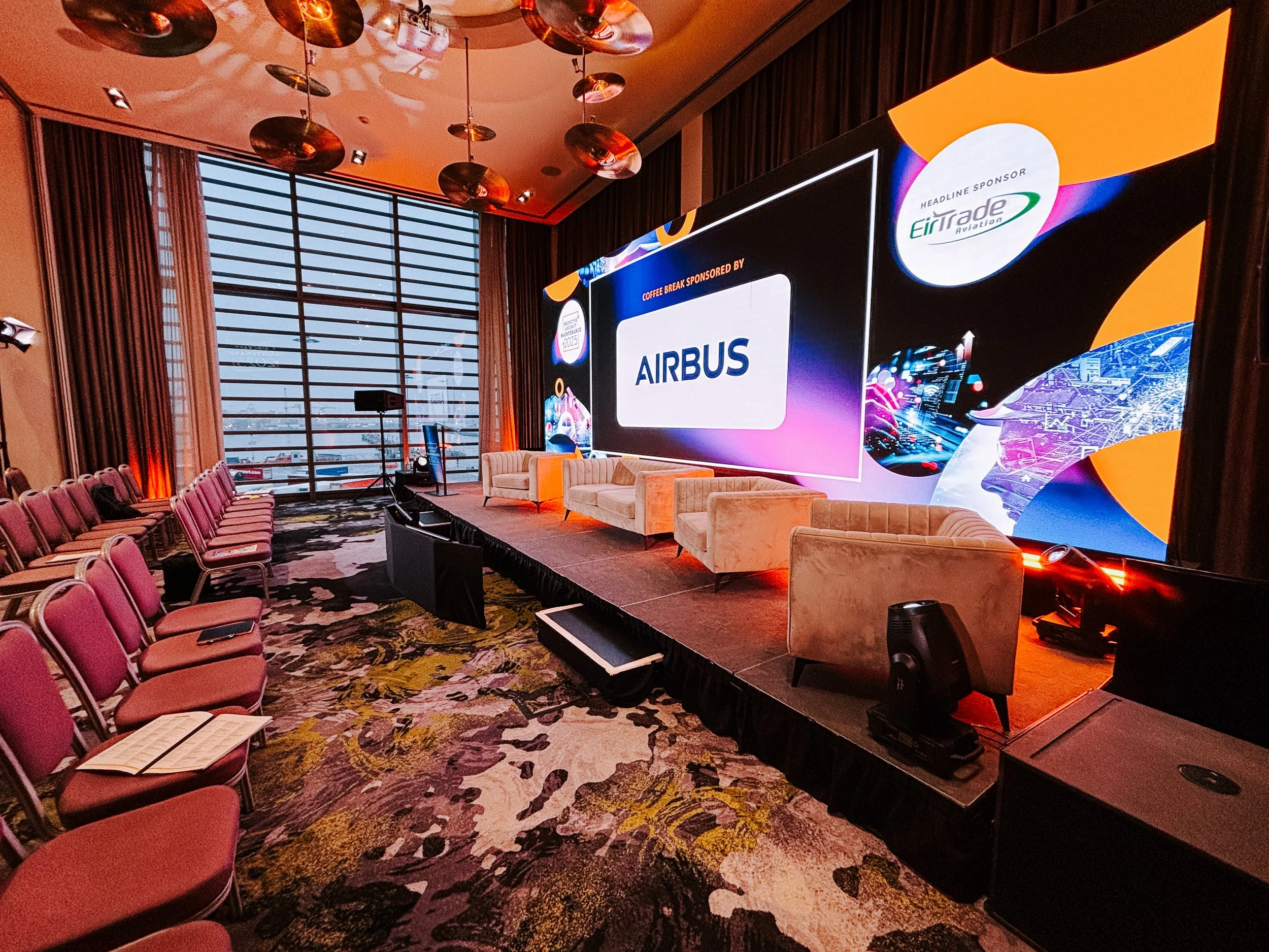 Conference room set up with rows of pink chairs facing a stage with beige chairs and a large screen displaying the Airbus logo and sponsor details, large windows with curtains, and modern lighting fixtures.