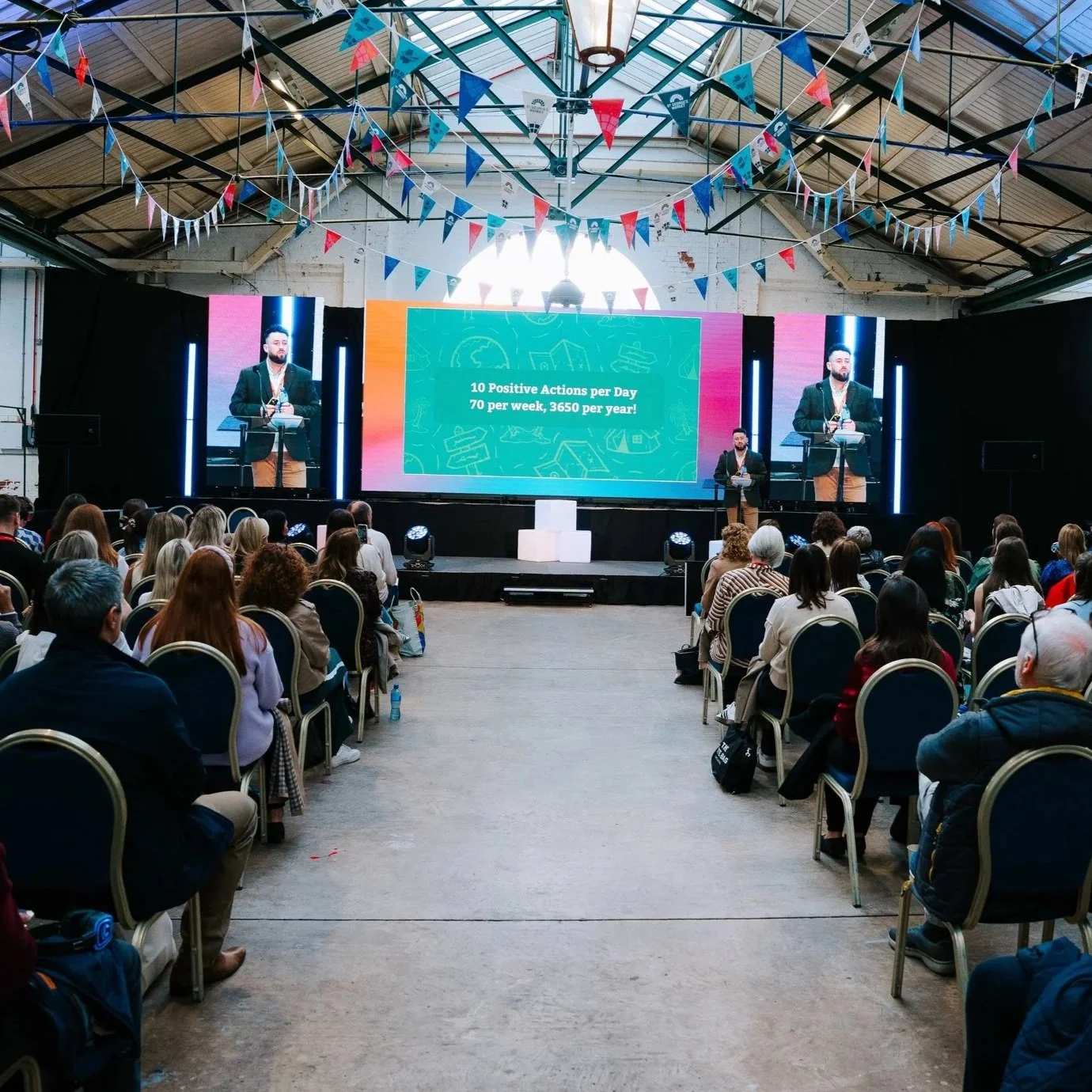Large conference or seminar event in a spacious industrial-style venue with a high ceiling, decorated with colorful triangular flags hanging from above. A speaker stands on stage delivering a presentation, with a large screen behind displaying a slide that reads "10 Positive Actions per Day, 70 per week, 3650 per year!". The audience sits in rows of chairs, attentively listening to the presentation.