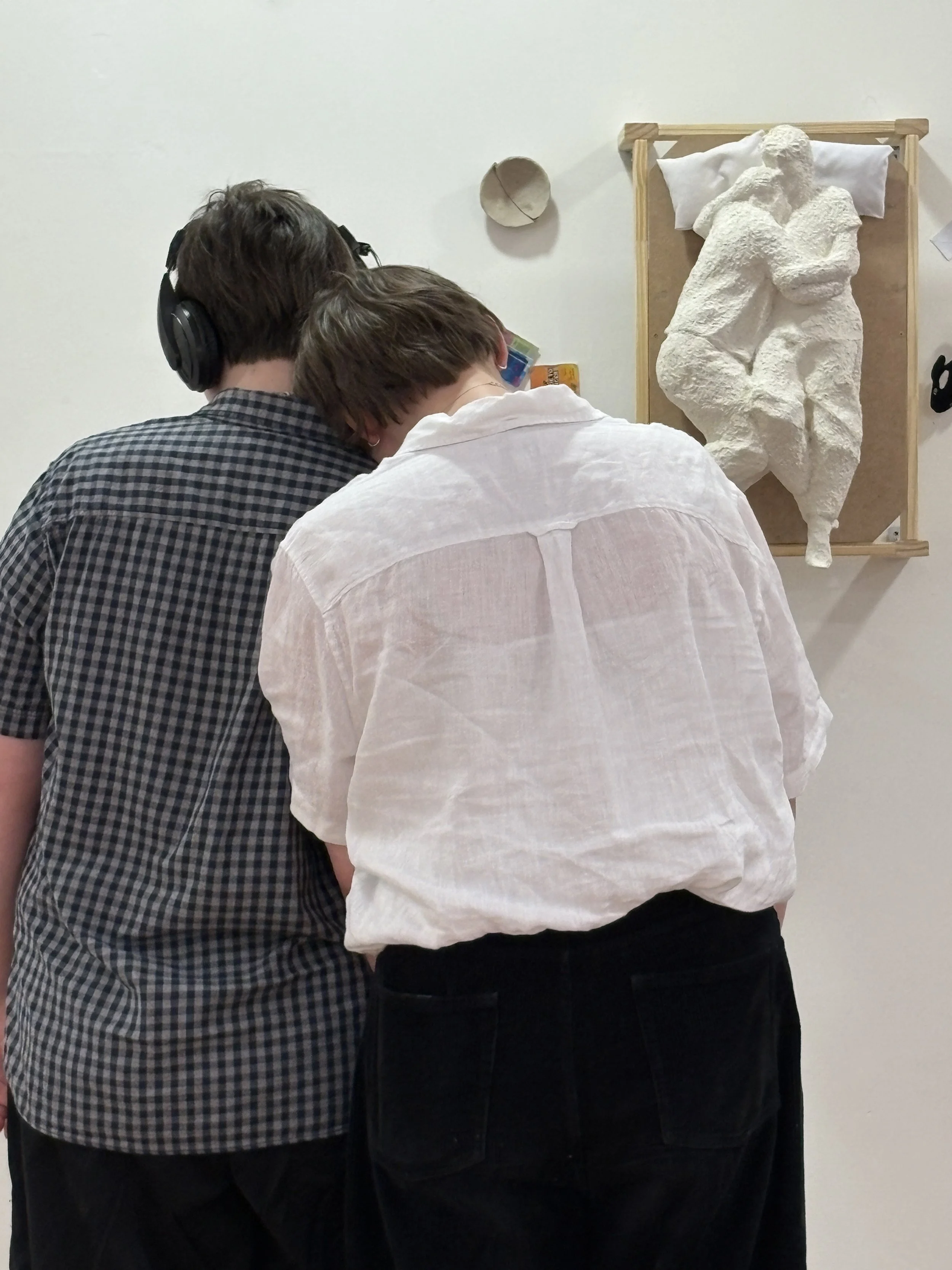 Two people, one with brown hair and plaid shirt and the other with short hair and white shirt, are standing close together with their backs to the camera, looking at an art sculpture on the wall that depicts two figures embracing.