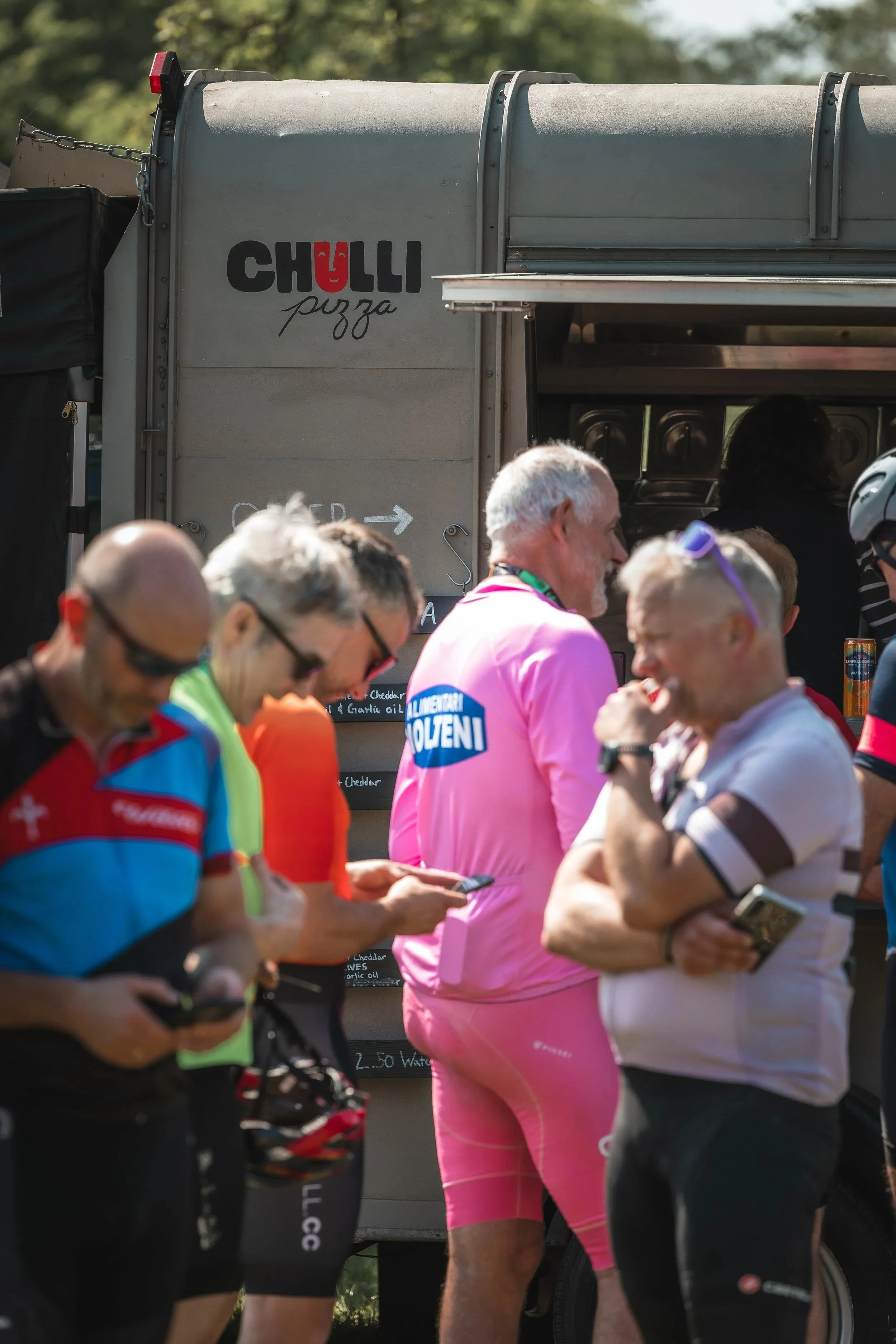 Group of cyclists with one person in pink riding gear, standing in front of a food truck called Chulli Pizza, with people checking their phones.