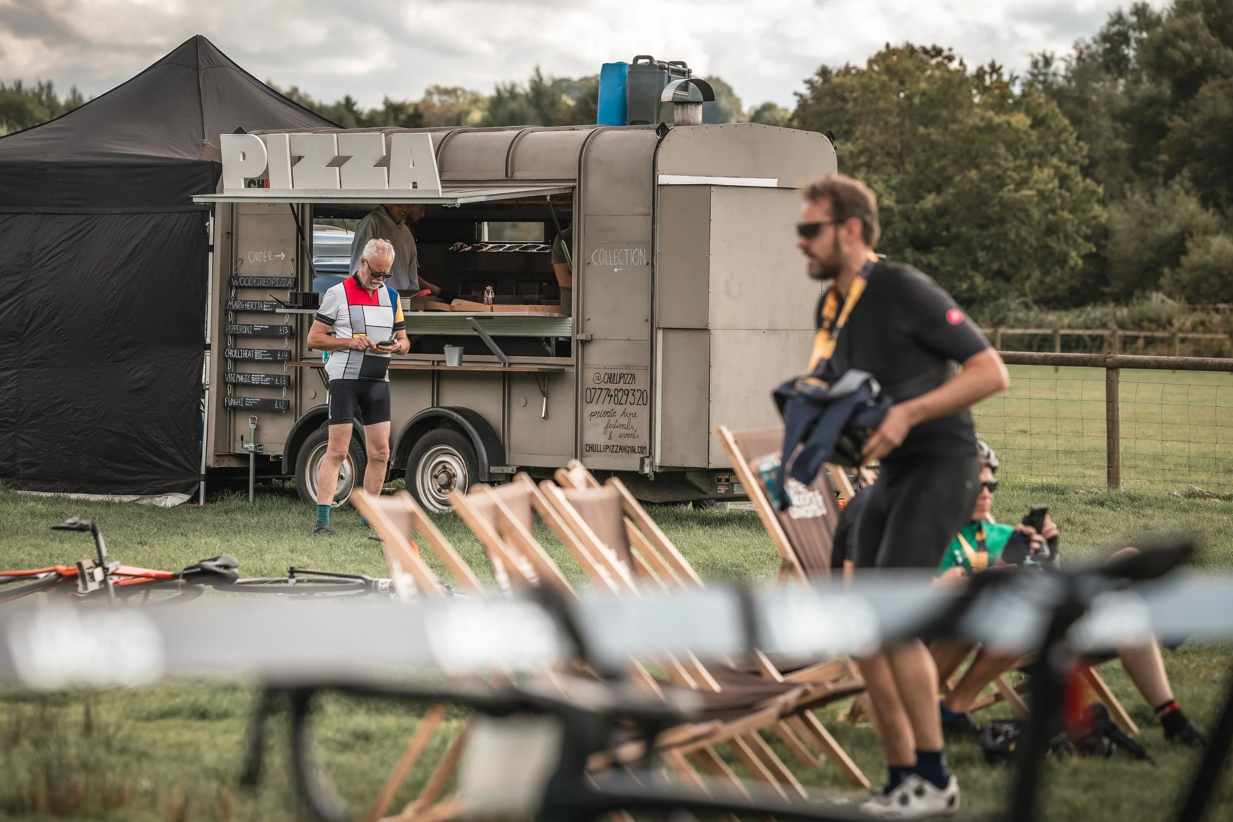 A food truck with a sign that reads 'PIZZA' parked outdoors next to a black tent, with a man in a cycling jersey looking at his phone in front of it. Several bicycles and outdoor chairs are scattered around, and there are trees in the background.