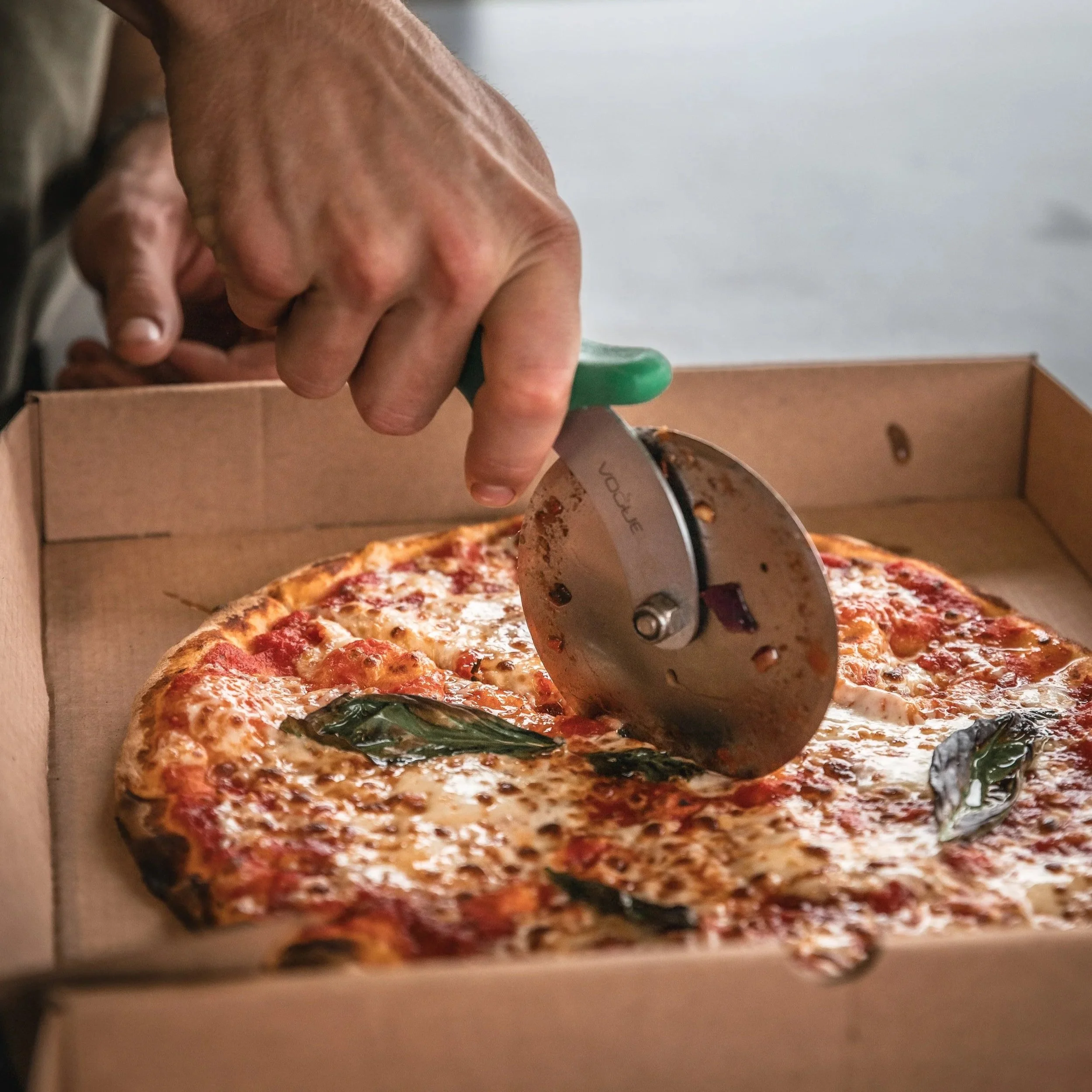 A pizza inside a cardboard box with a hand cutting the pizza with a pizza cutter.
