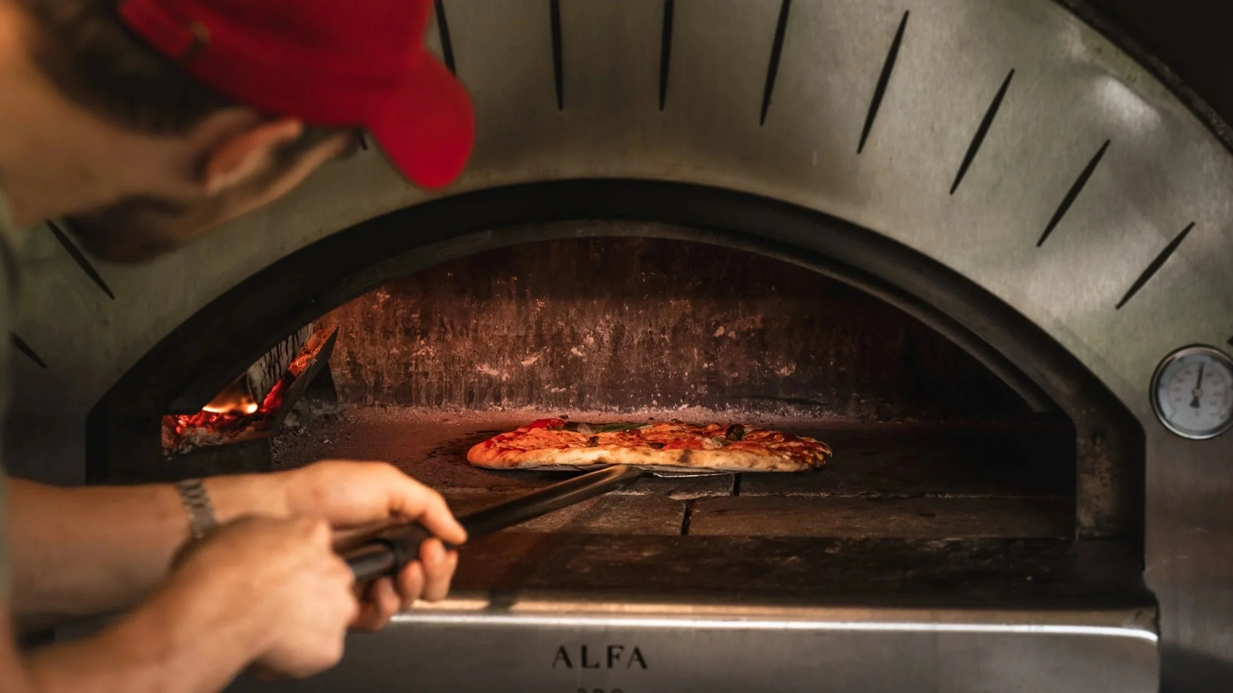 Person placing pizza into an industrial pizza oven inside of the chulli pizza trailer