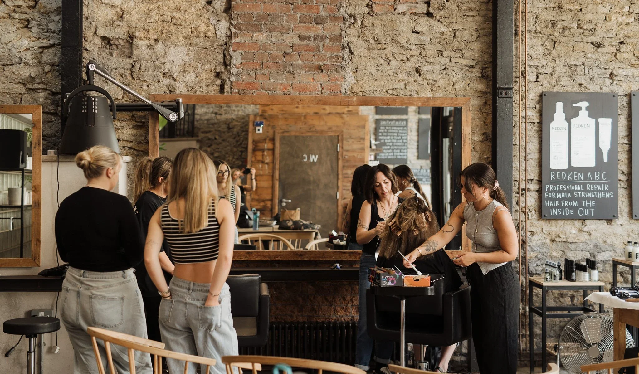 A group of women gathers in a hair salon. One woman is getting her hair styled or colored by a stylist, while others wait or observe. The salon has a rustic brick wall, a mirror, and shelves with hair products. There is a sign advertising Redken hair products and treatments.