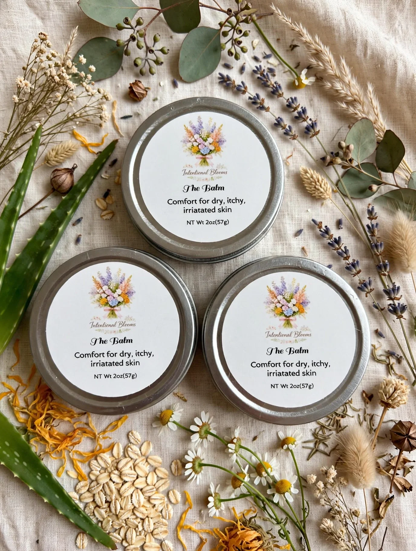 Three metal tins labeled 'The Balm,' with floral illustrations and text indicating they are for dry, itchy, irritated skin, placed on a linen surface decorated with various dried flowers and greenery.